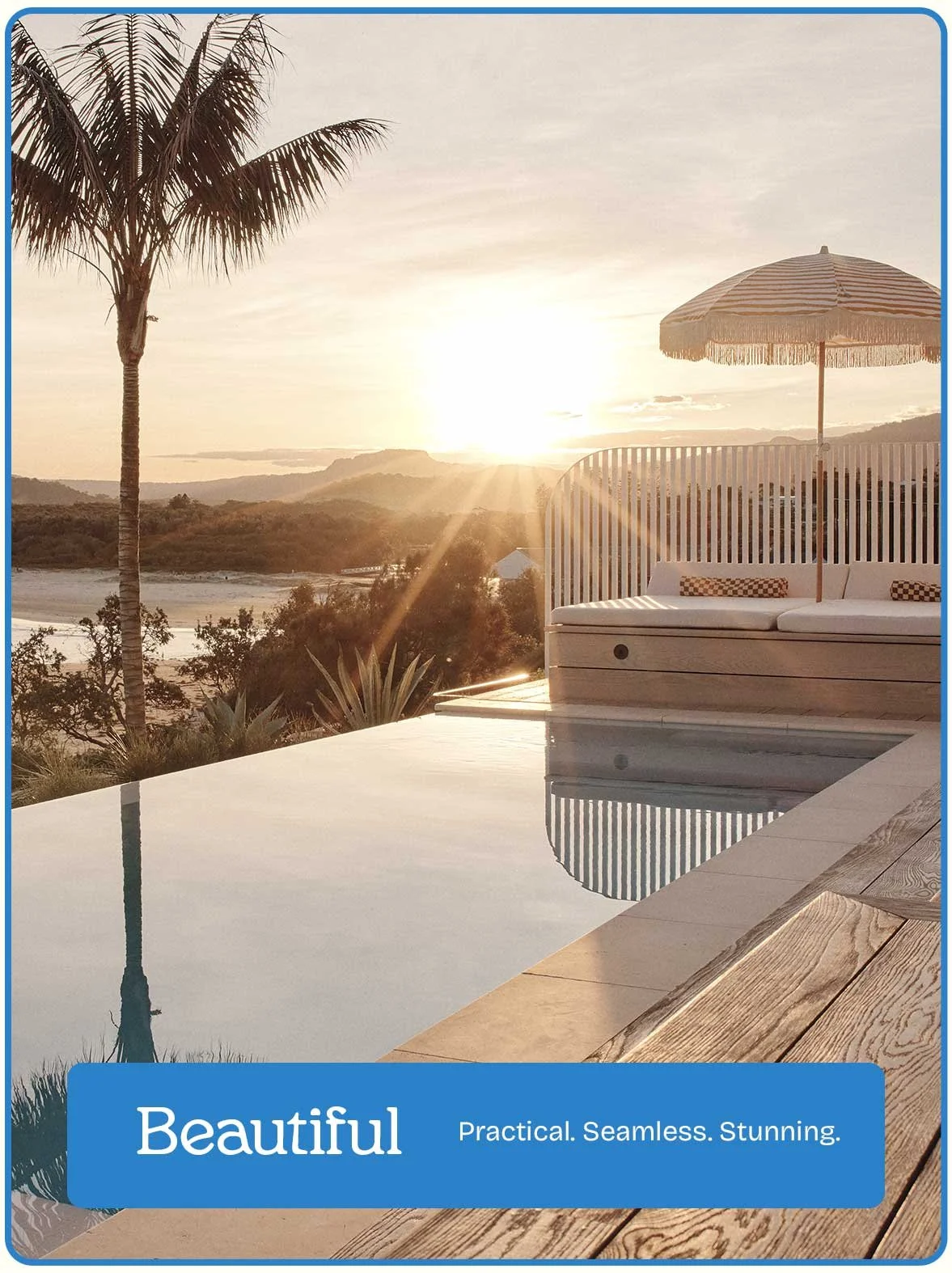 A scenic outdoor view during sunset with a palm tree, a swimming pool, a wooden deck, and a sun umbrella on a deck chair, overlooking distant hills and a river.