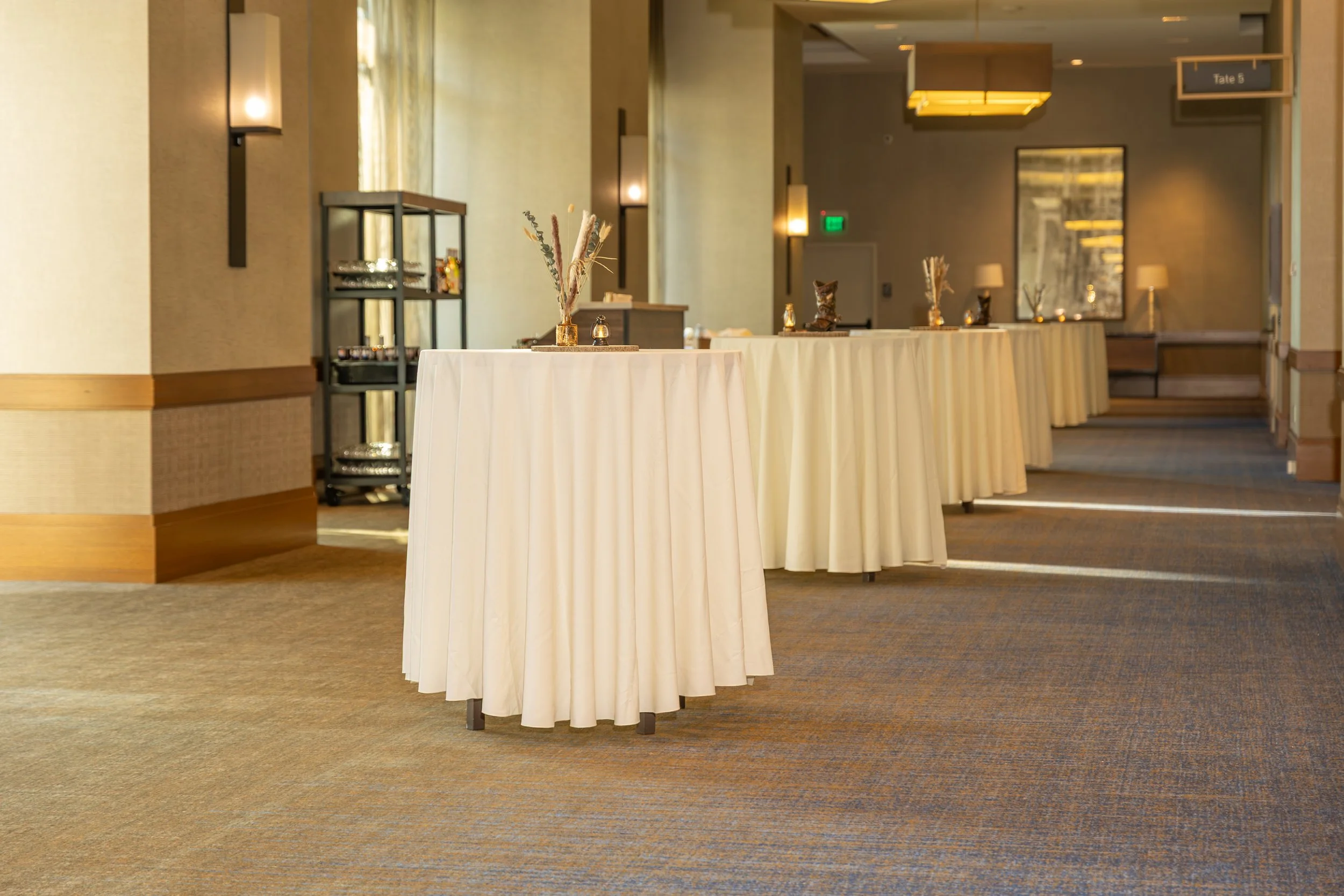Elegant hotel event space with white cocktail tables, floral arrangements, warm lighting, and a Tate B meeting room sign