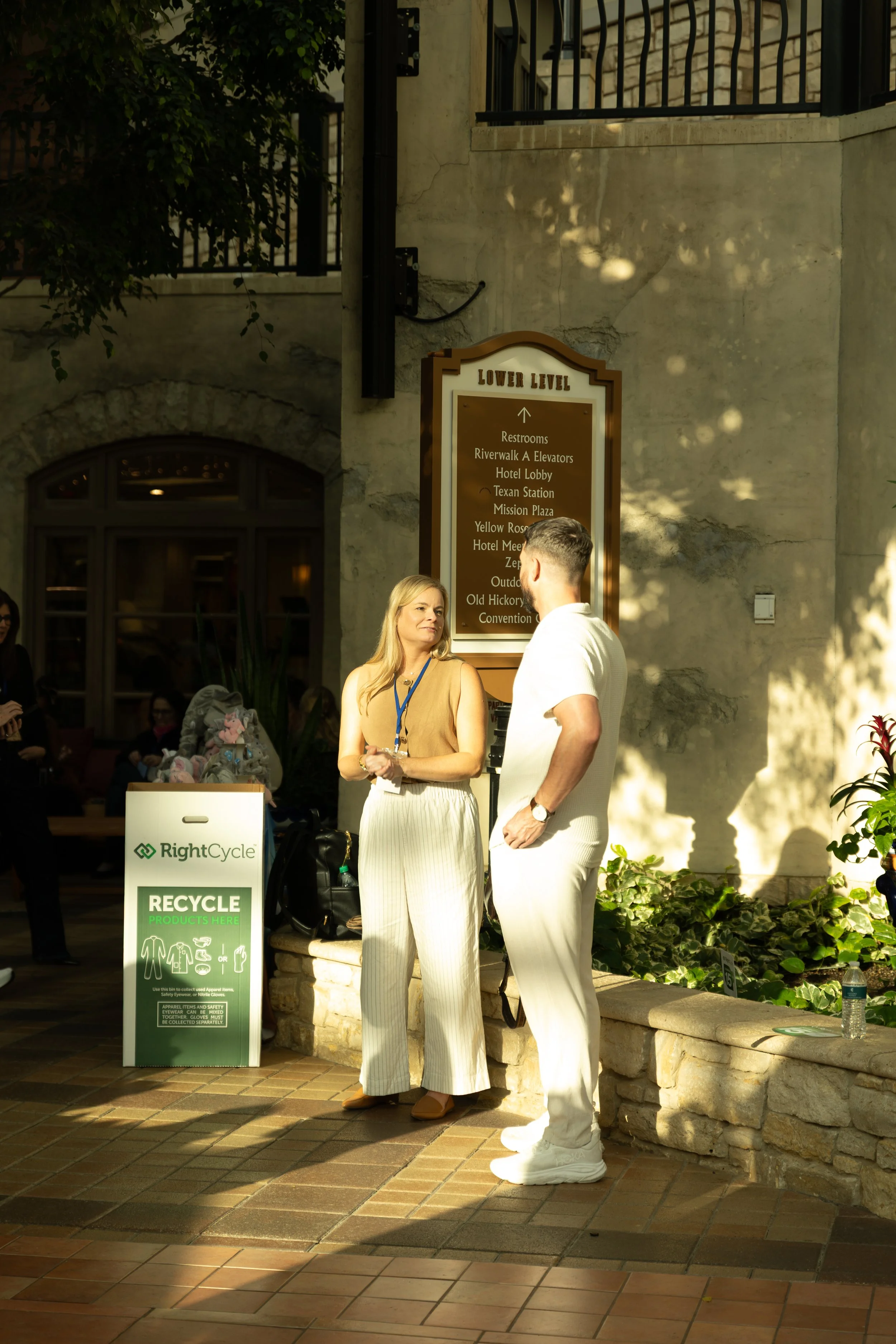Two people stand beside a lower-level directional sign and RightCycle recycling bin in a sunlit hotel lobby.
