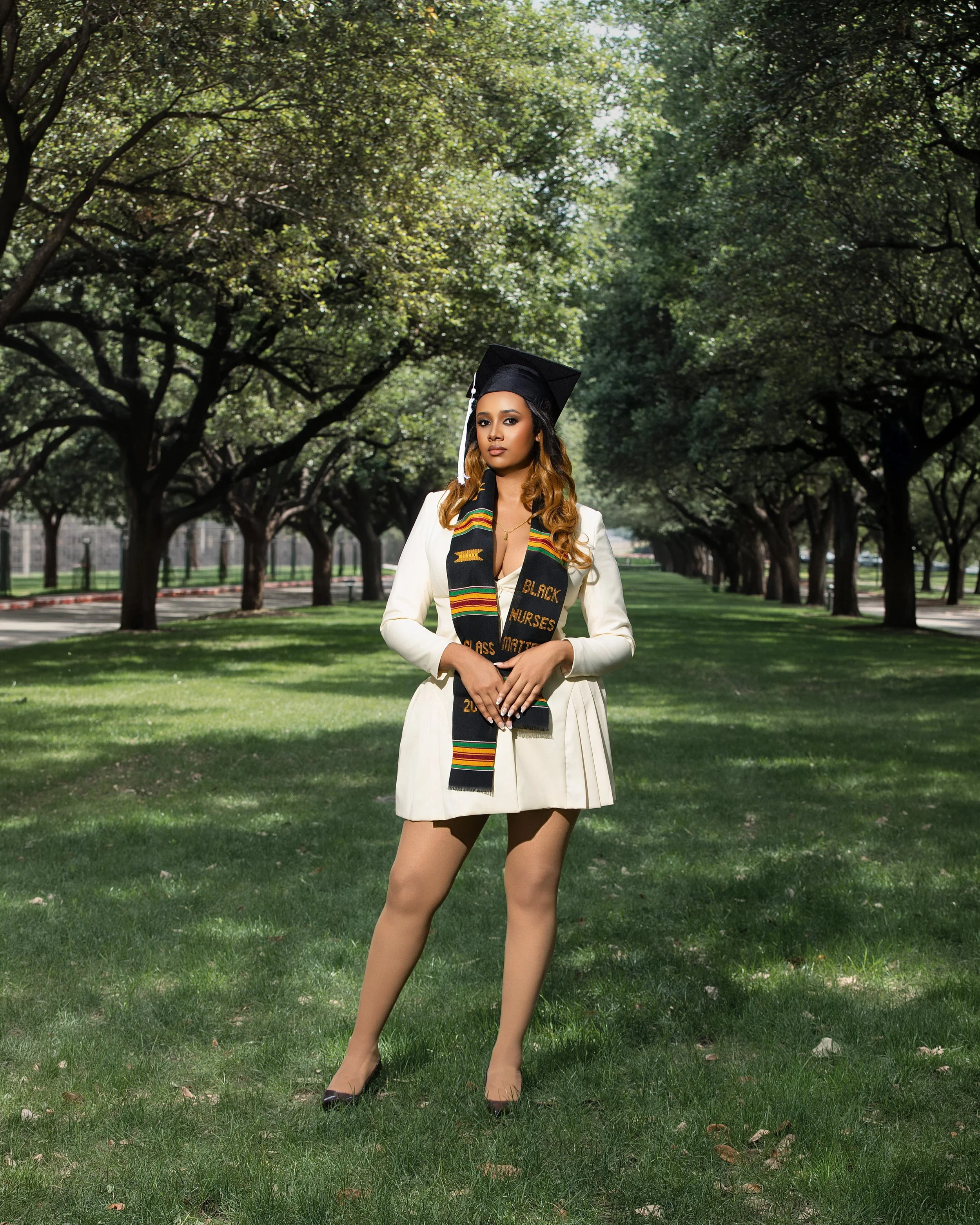 A woman in a white graduation gown and cap stands outdoors on grass, holding a stole that says "Black Nurses Matter," with trees and a park path in the background.