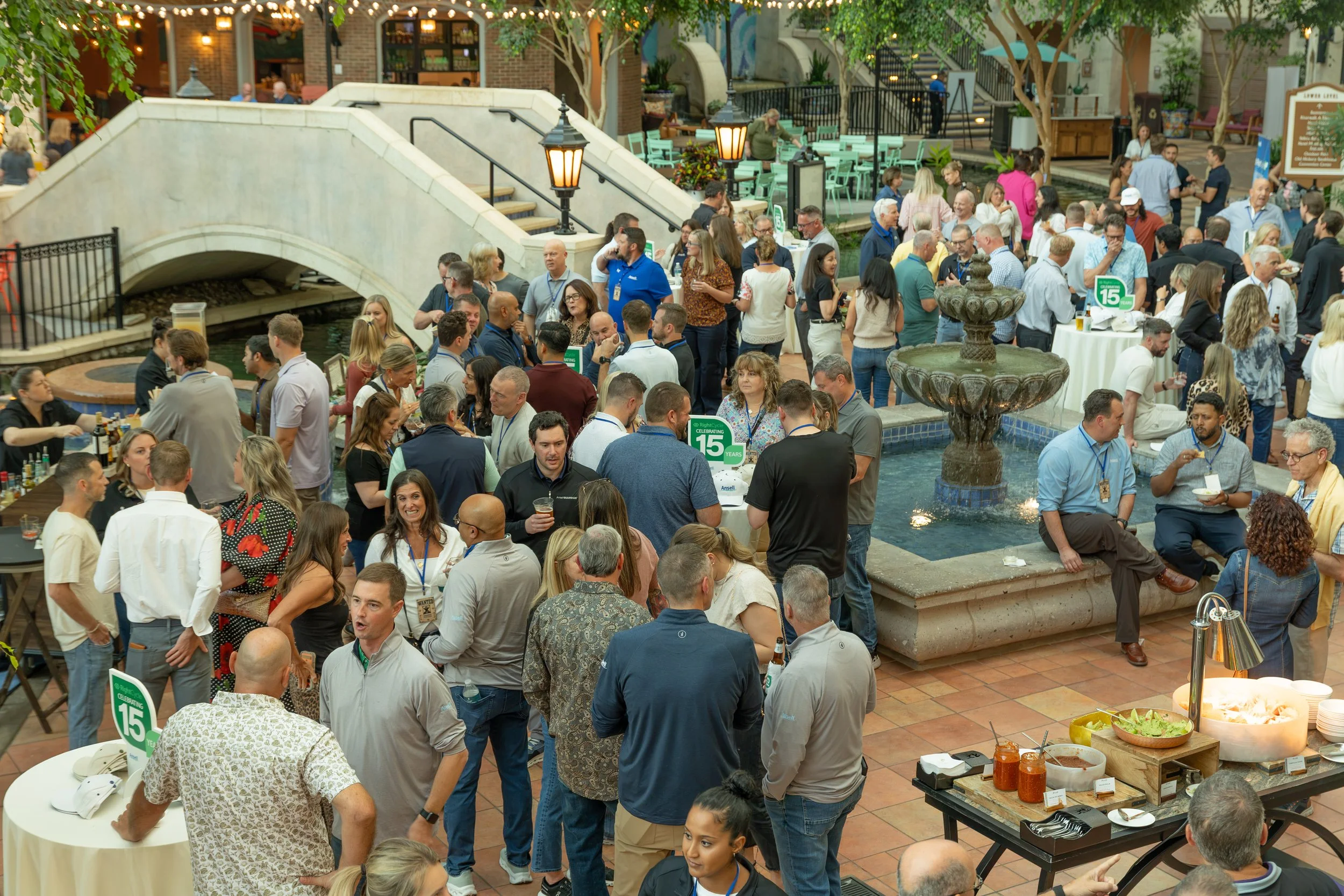 Crowded indoor networking event with guests, buffet tables, fountain, bridge, and string lights in an atrium