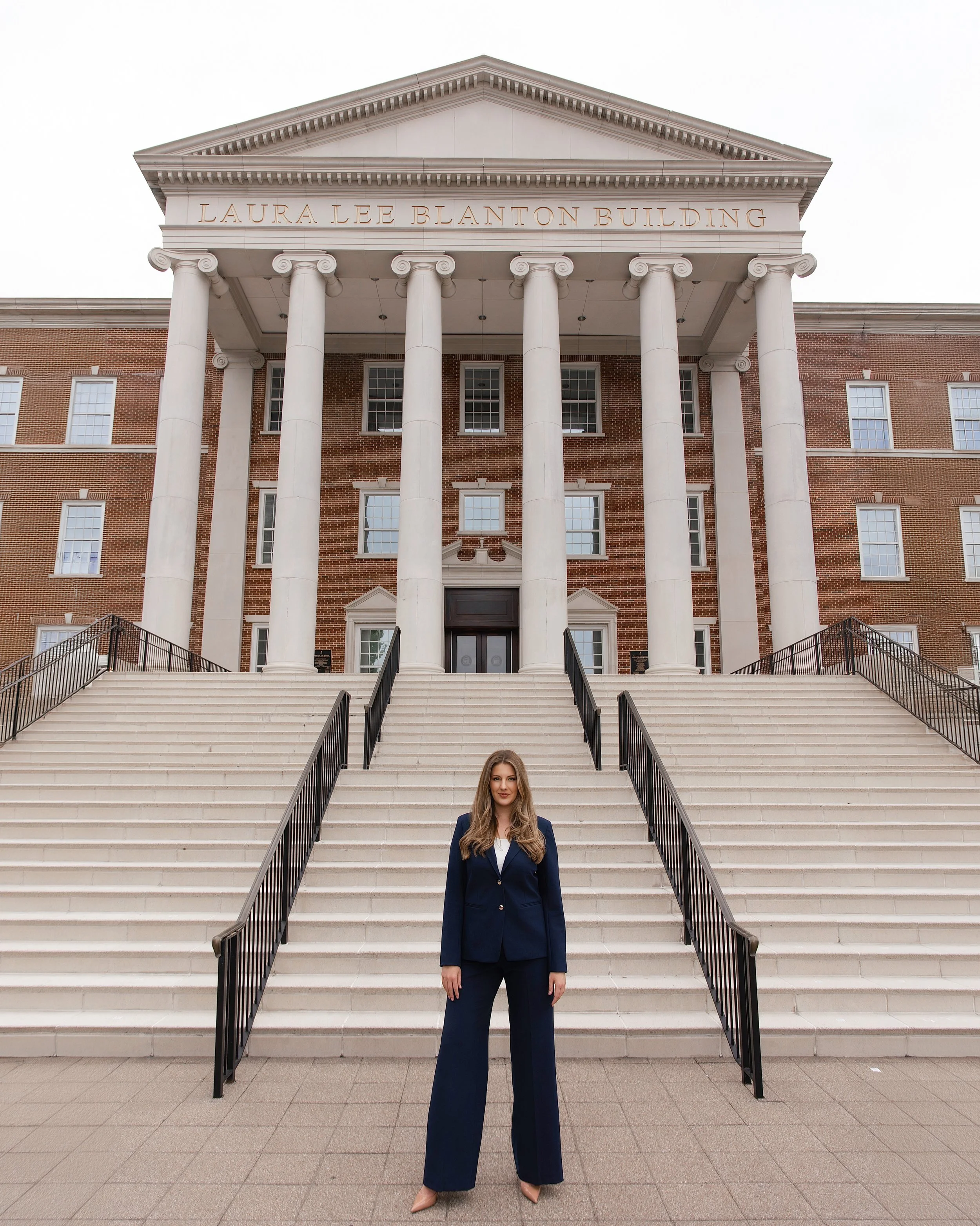 A woman with long blonde hair standing on steps leading to a large, neoclassical-style building with six tall white columns and the name 'Laura Lee Blanton Building' inscribed above the columns.