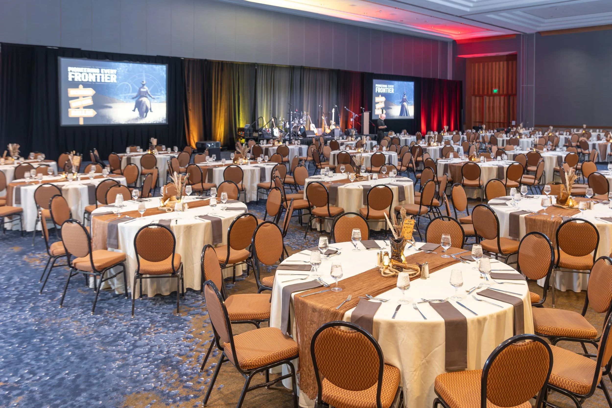 Elegant banquet hall with round dining tables, place settings, and a decorated stage with musical instruments and projection screens