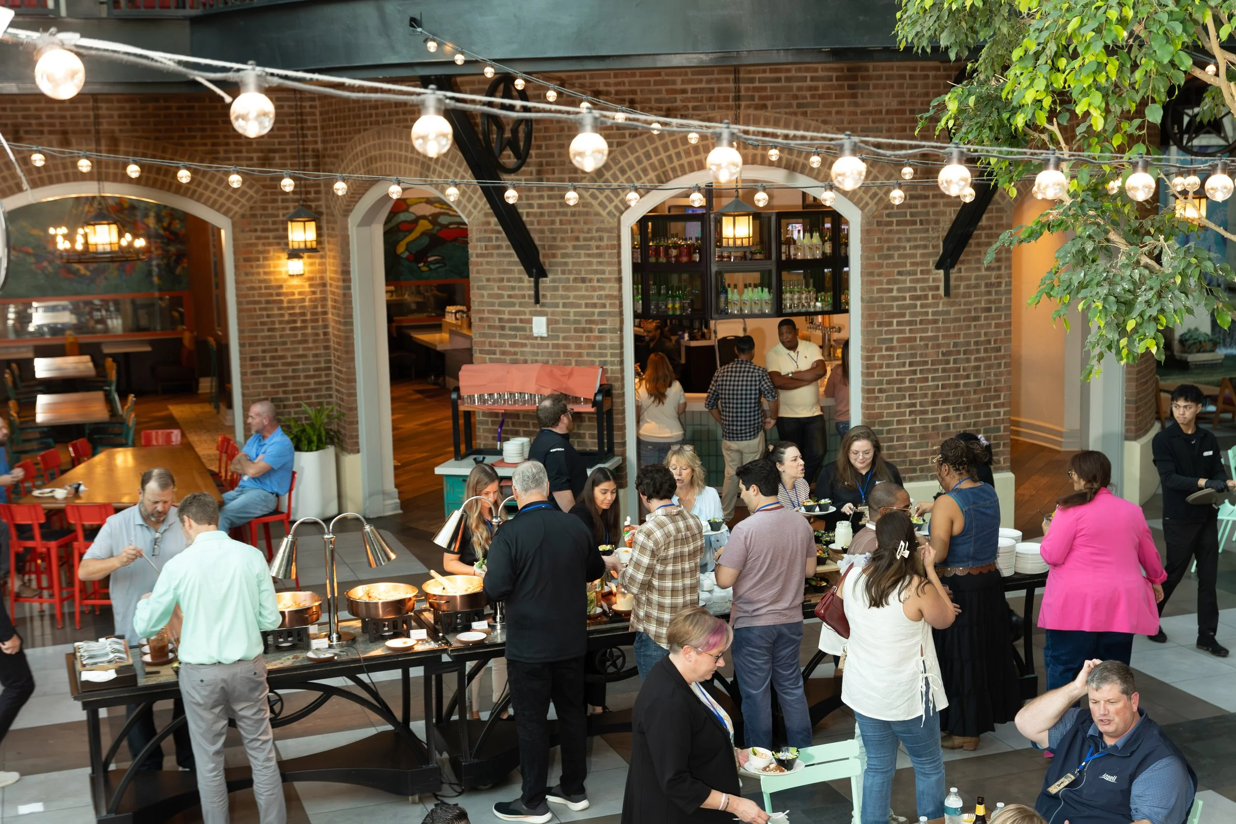 Busy restaurant buffet with diners, brick arches, a bar, and hanging string lights