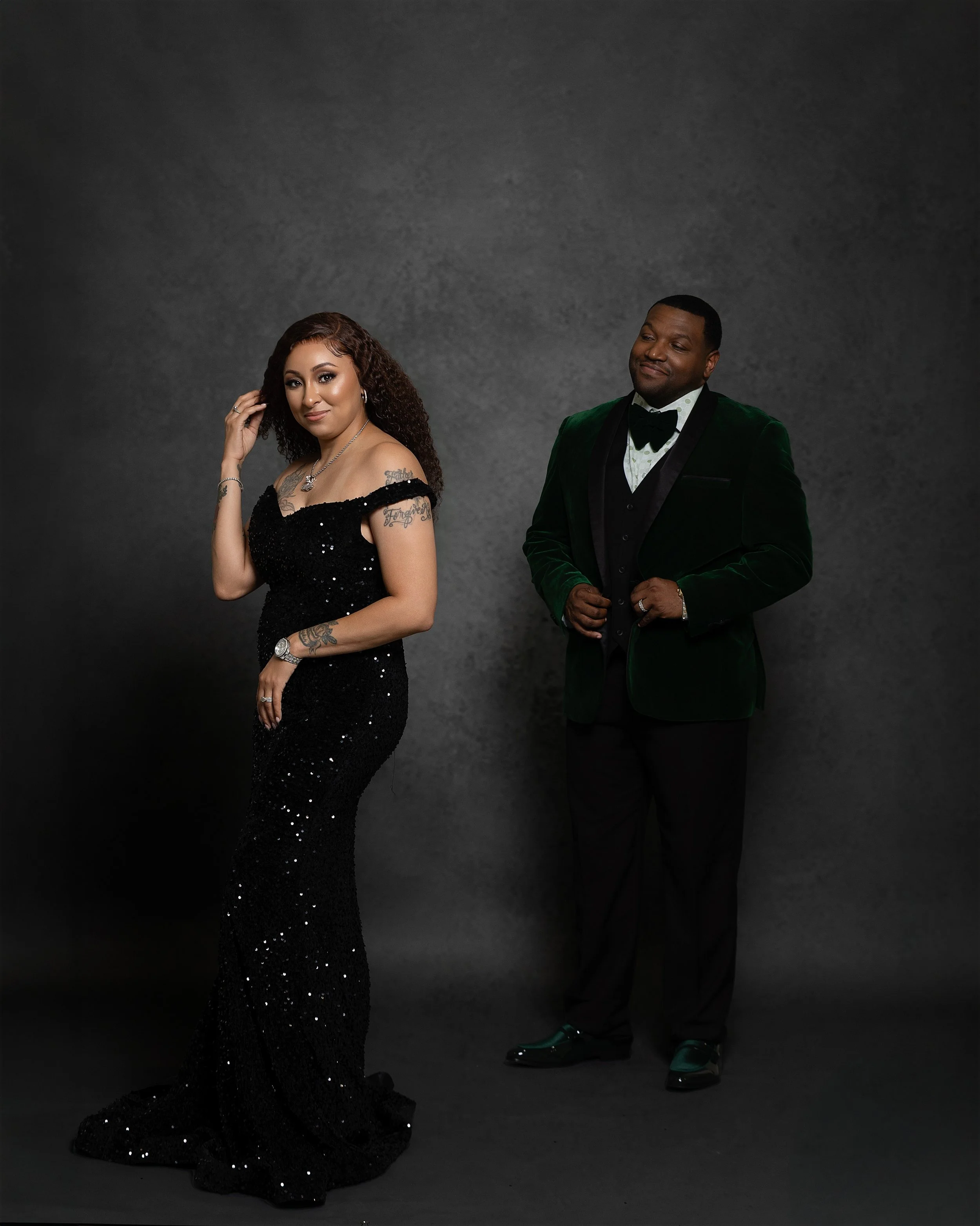 A woman in a strapless black dress with sequins and tattoos on her arms, and a man in a dark green velvet tuxedo with black lapels, white shirt, and black bow tie, standing against a plain gray background.