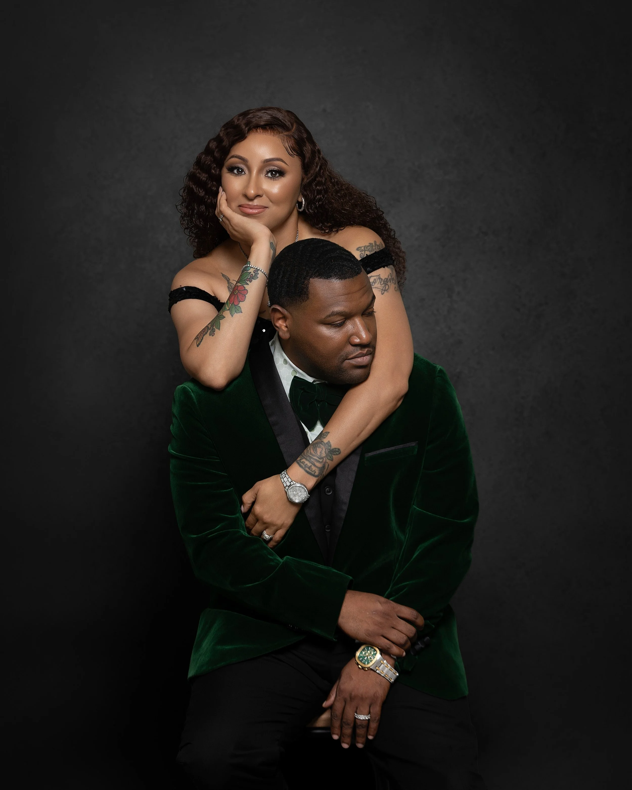 A woman with curly hair and tattoos on her arms is sitting on a man's lap. The man is wearing a dark green velvet blazer and a white shirt. The woman is resting her face on her hand, and both appear relaxed against a dark background.