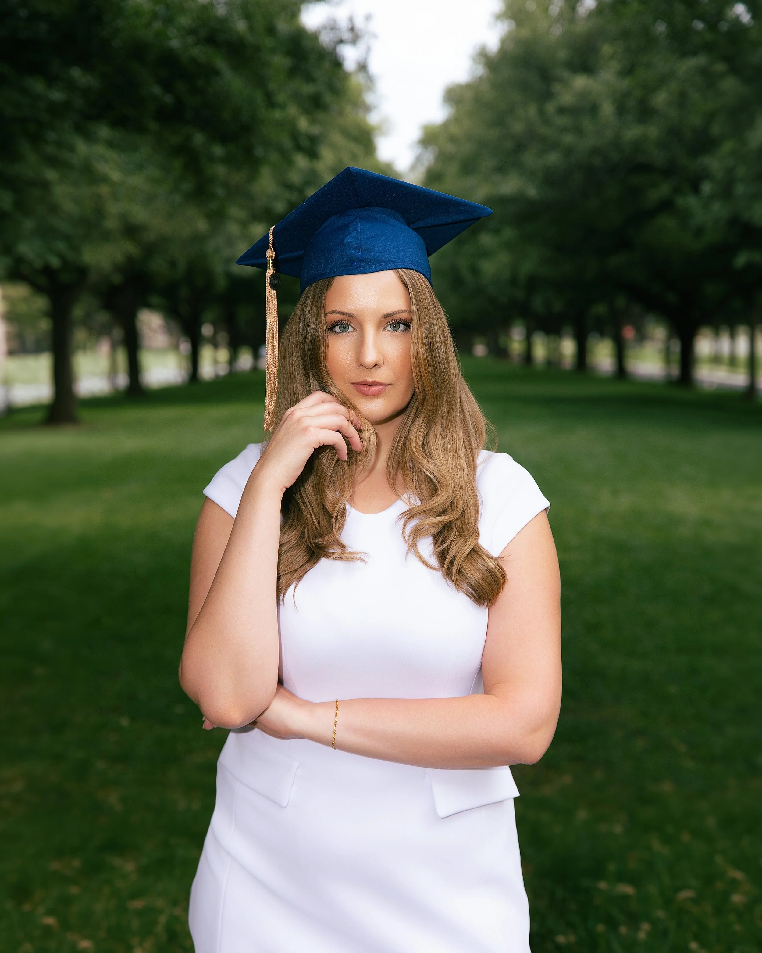 Young woman with long wavy hair wearing a white dress and a blue graduation cap, standing outdoors in a park with green trees and grass background.