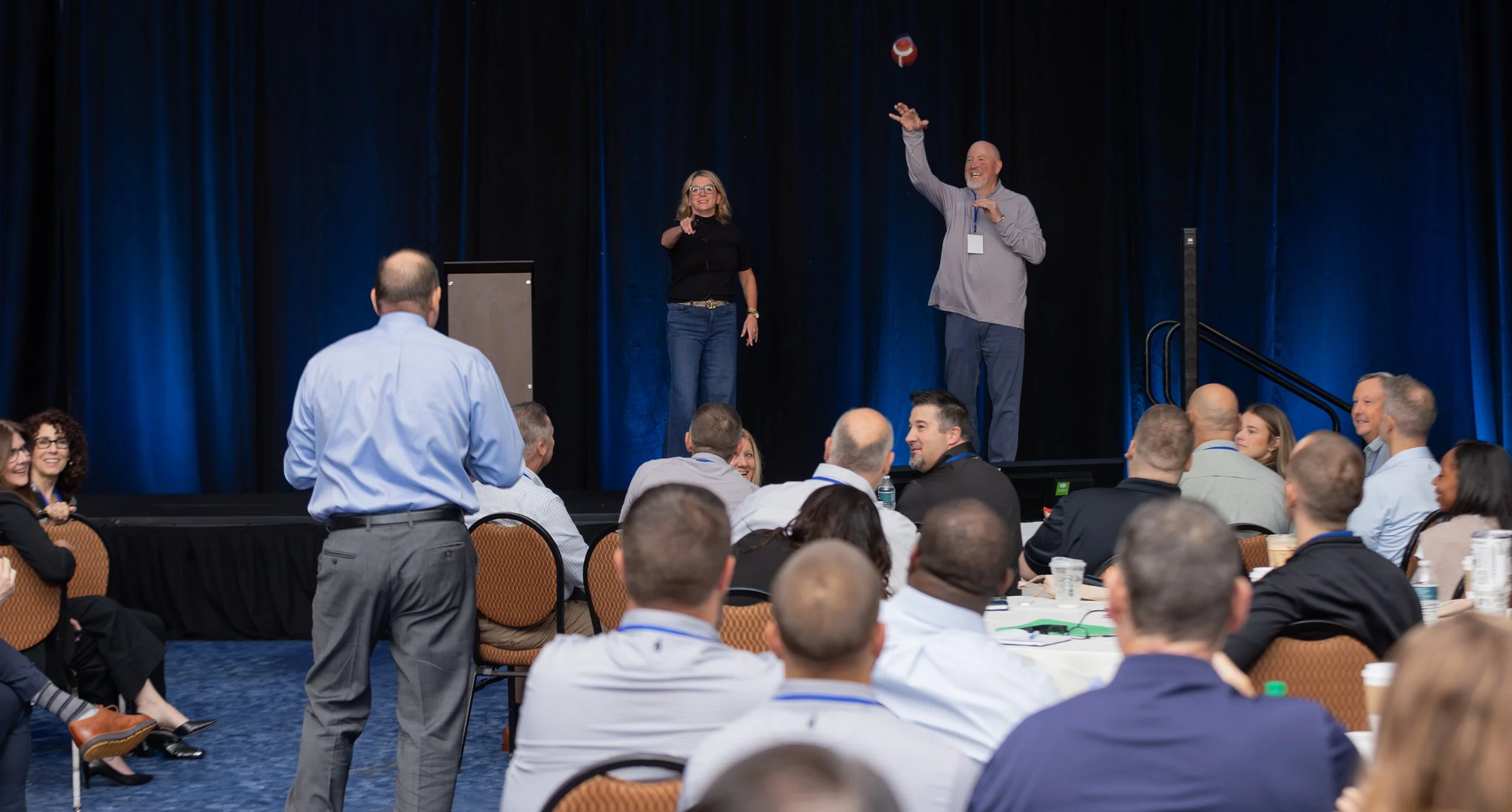 Conference audience watches two presenters onstage as a football arcs through the air