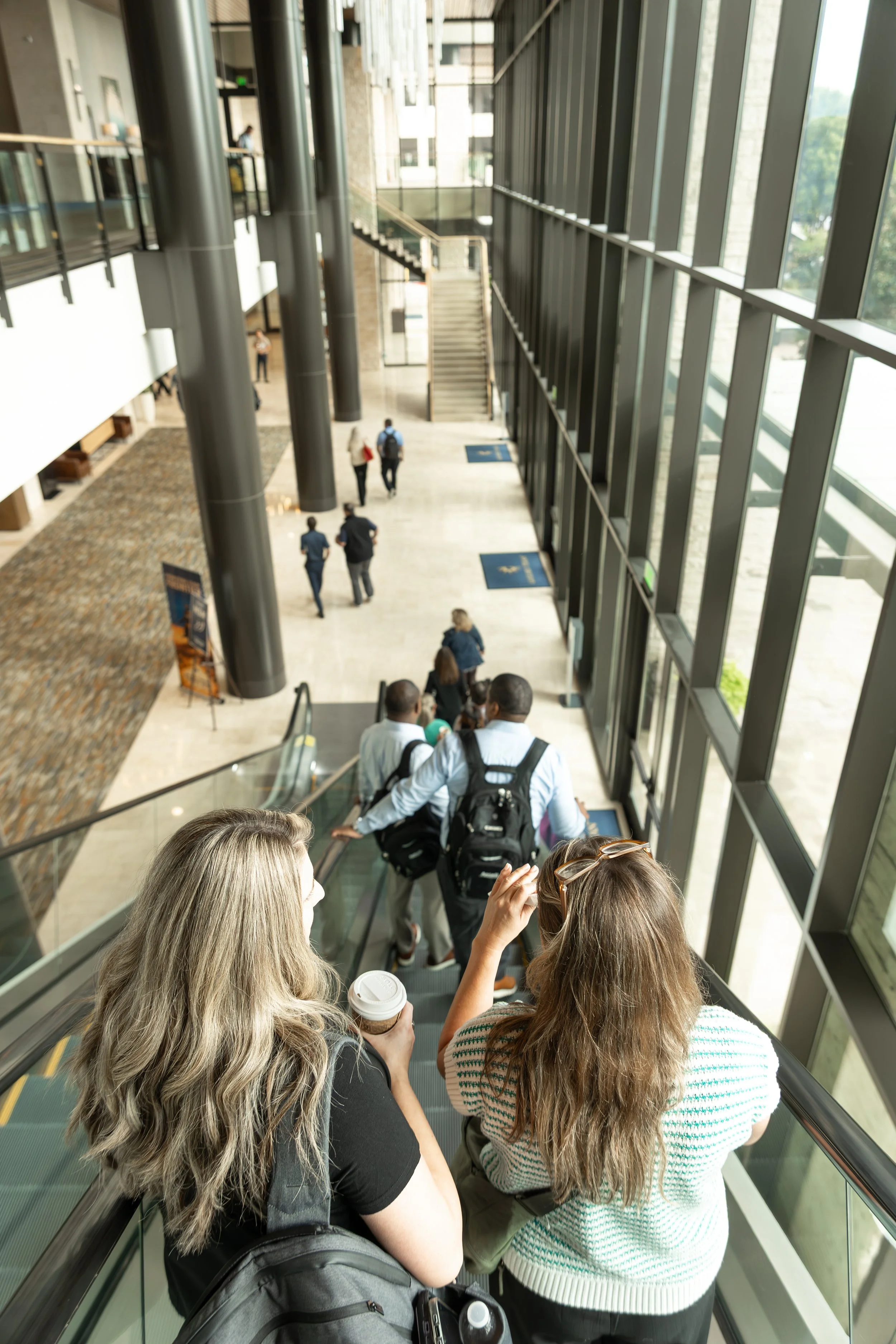 People descending an escalator inside a modern, sunlit building atrium with large windows, columns, and a staircase