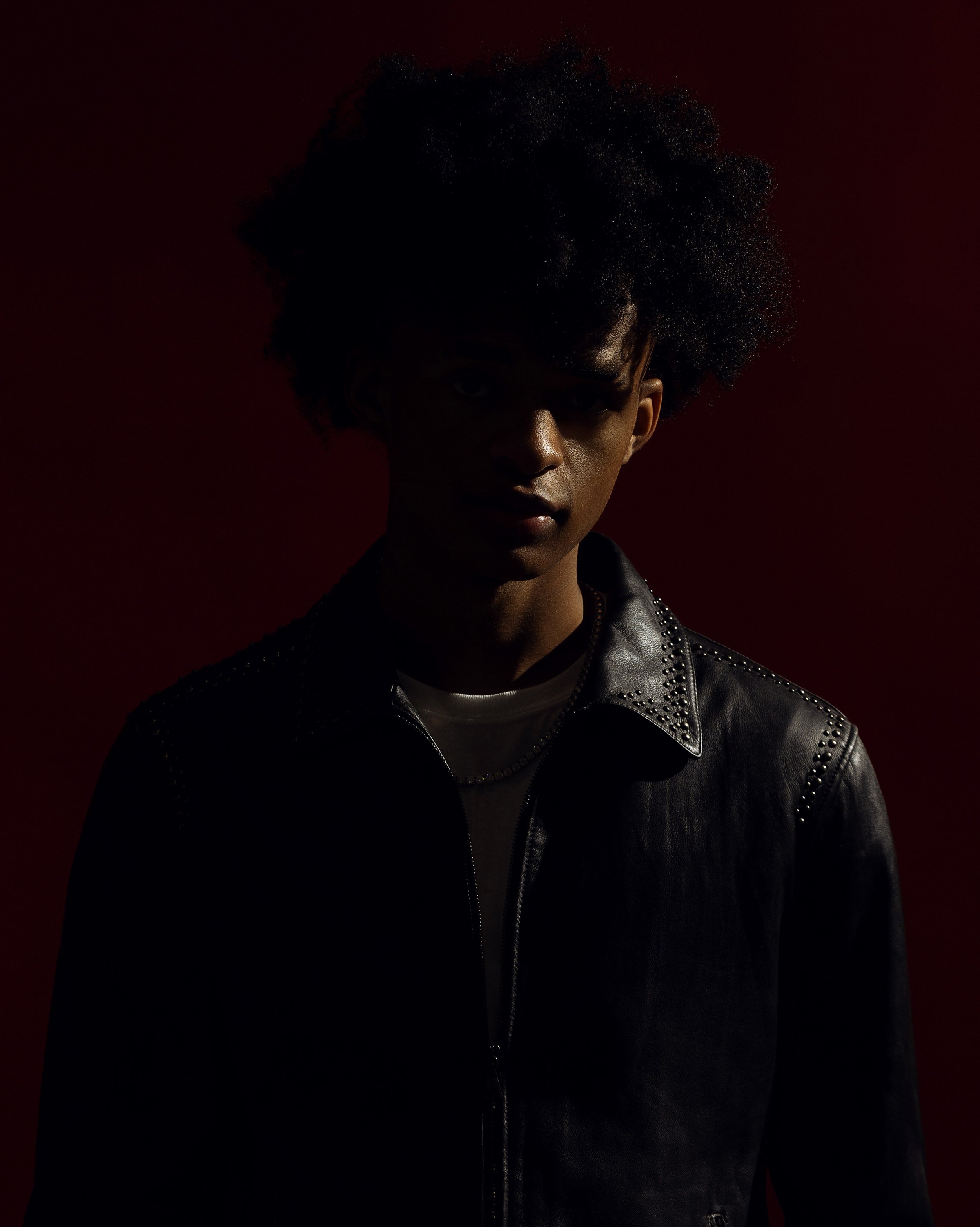 Person with an afro hairstyle wearing a black leather jacket with studs, standing against a dark red background.