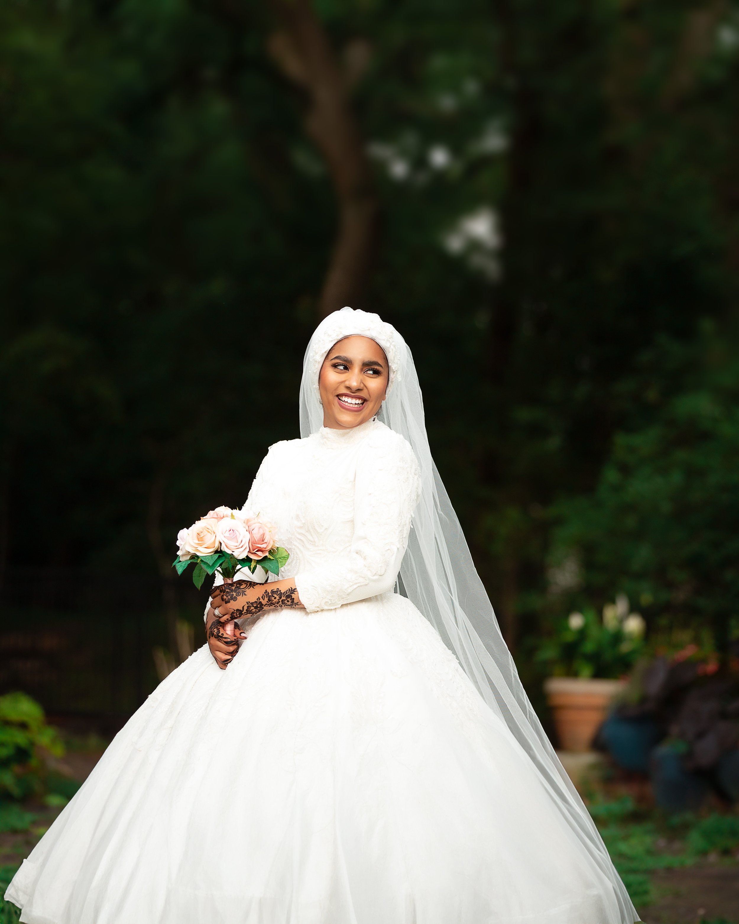 Bride in white wedding dress and veil holding bouquet, outdoor setting.