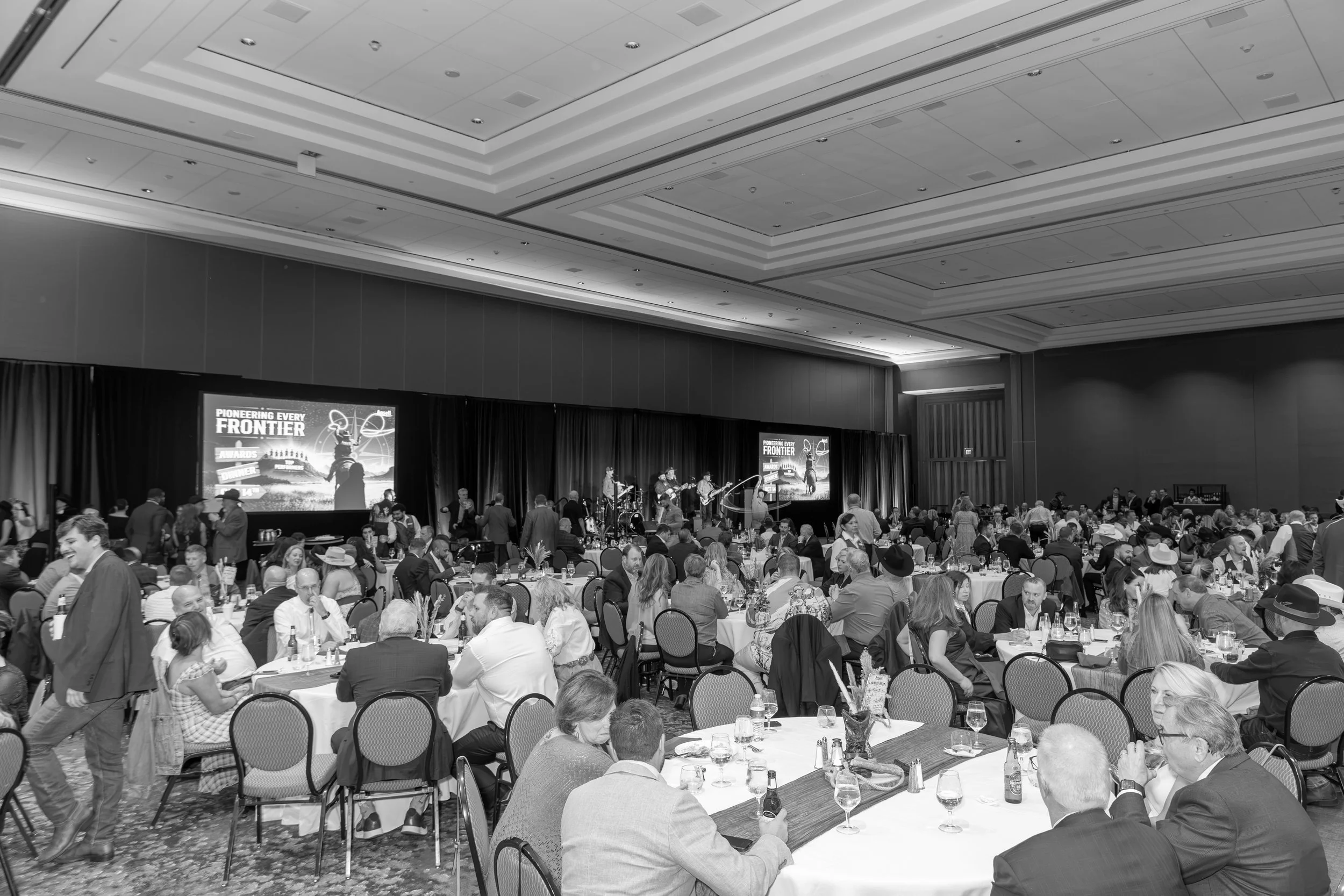 Large gala dinner with guests seated at round tables, a live band performing onstage, and screens displaying a Frontier awards event graphic.