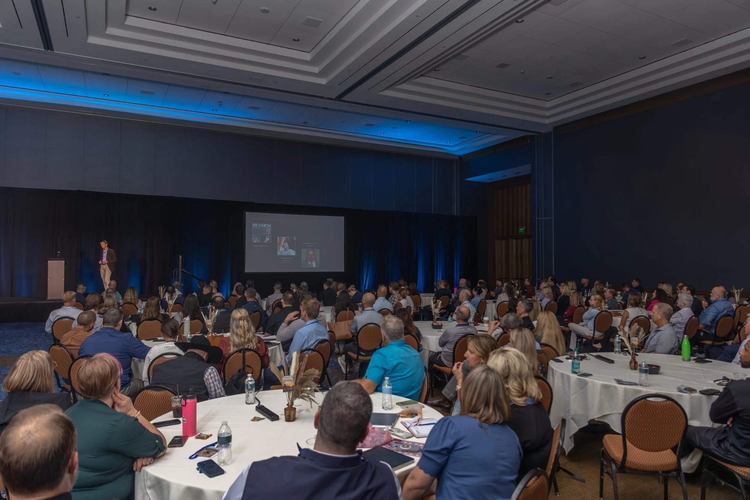 Speaker presenting to a large audience seated at round tables in a conference ballroom with blue lighting and a projection screen