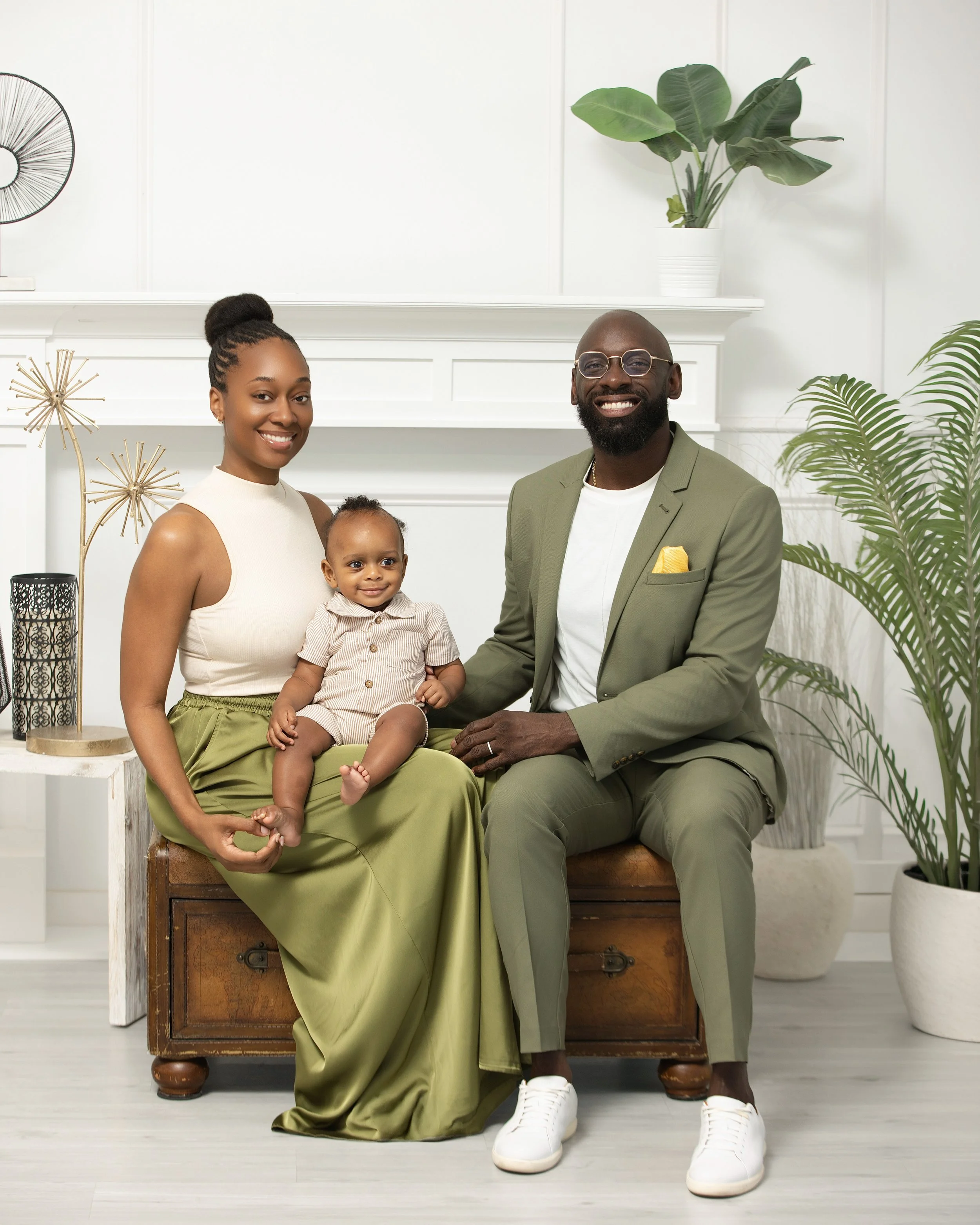 A family portrait with a woman, a man, and a young child sitting on a wooden trunk in a bright, stylish room with plants and modern decor.