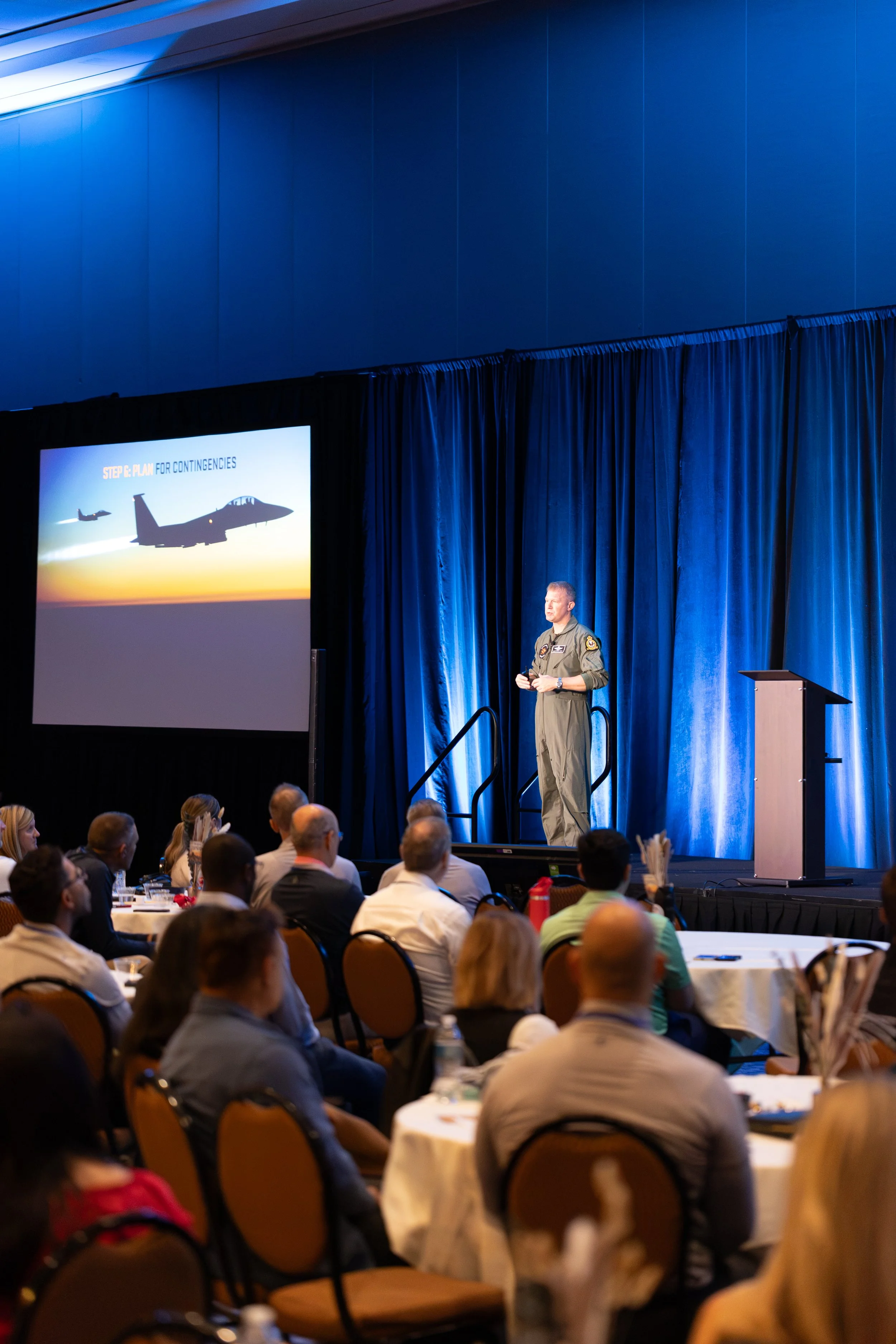 Military pilot speaking to an audience beside a presentation slide showing fighter jets on a blue-lit stage.