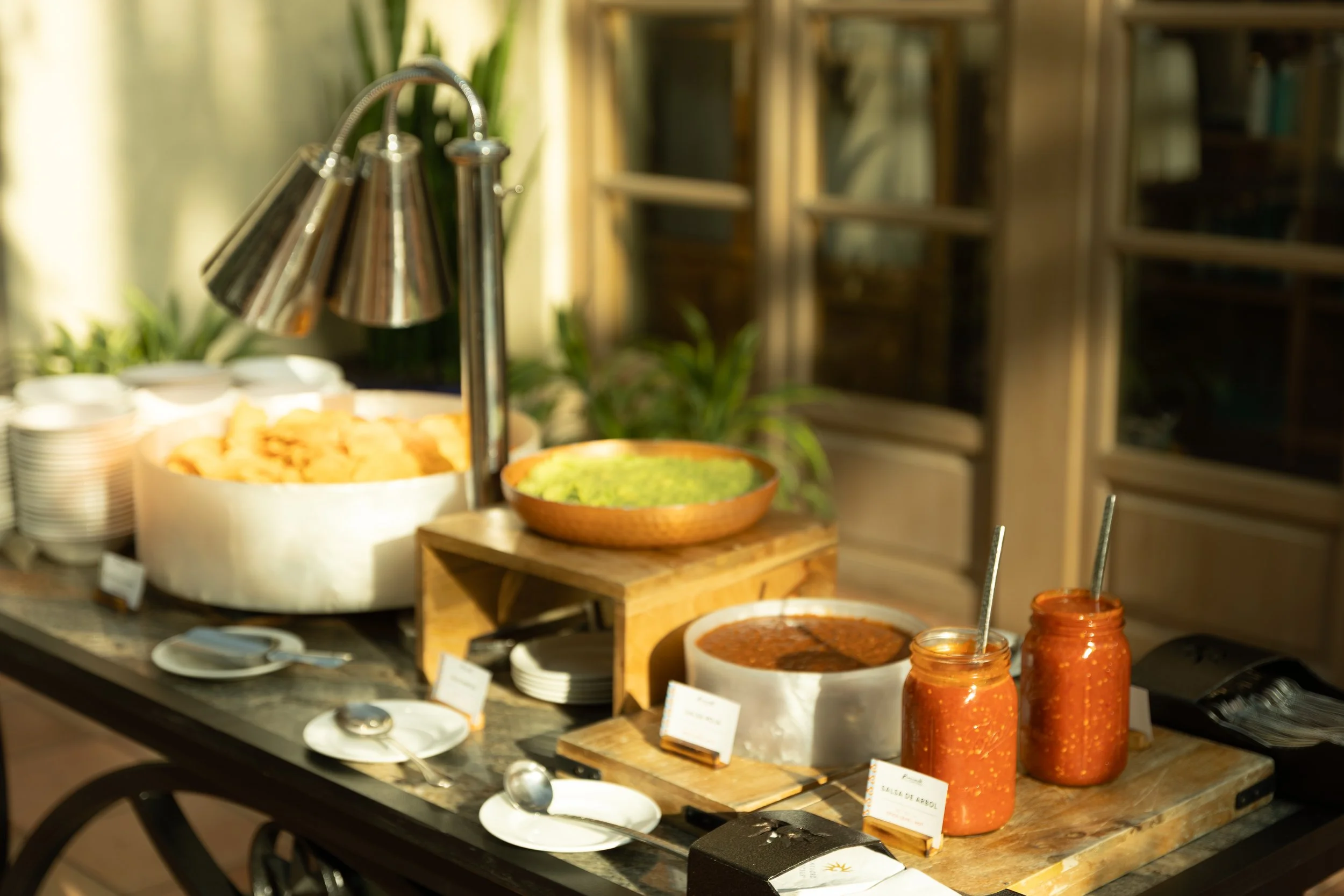 Buffet station with stainless steel serving utensils, bowls of dips and sauces, chips, plates, and serving spoons.