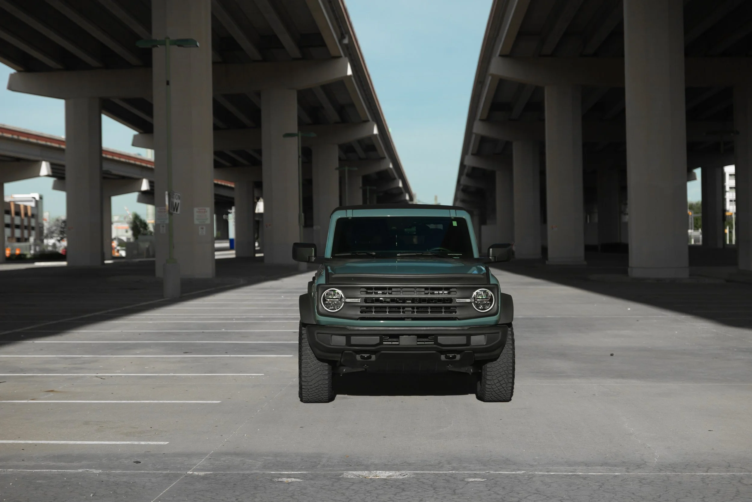 A green compact car parked alone in an empty parking lot underneath large elevated highways in an urban setting.