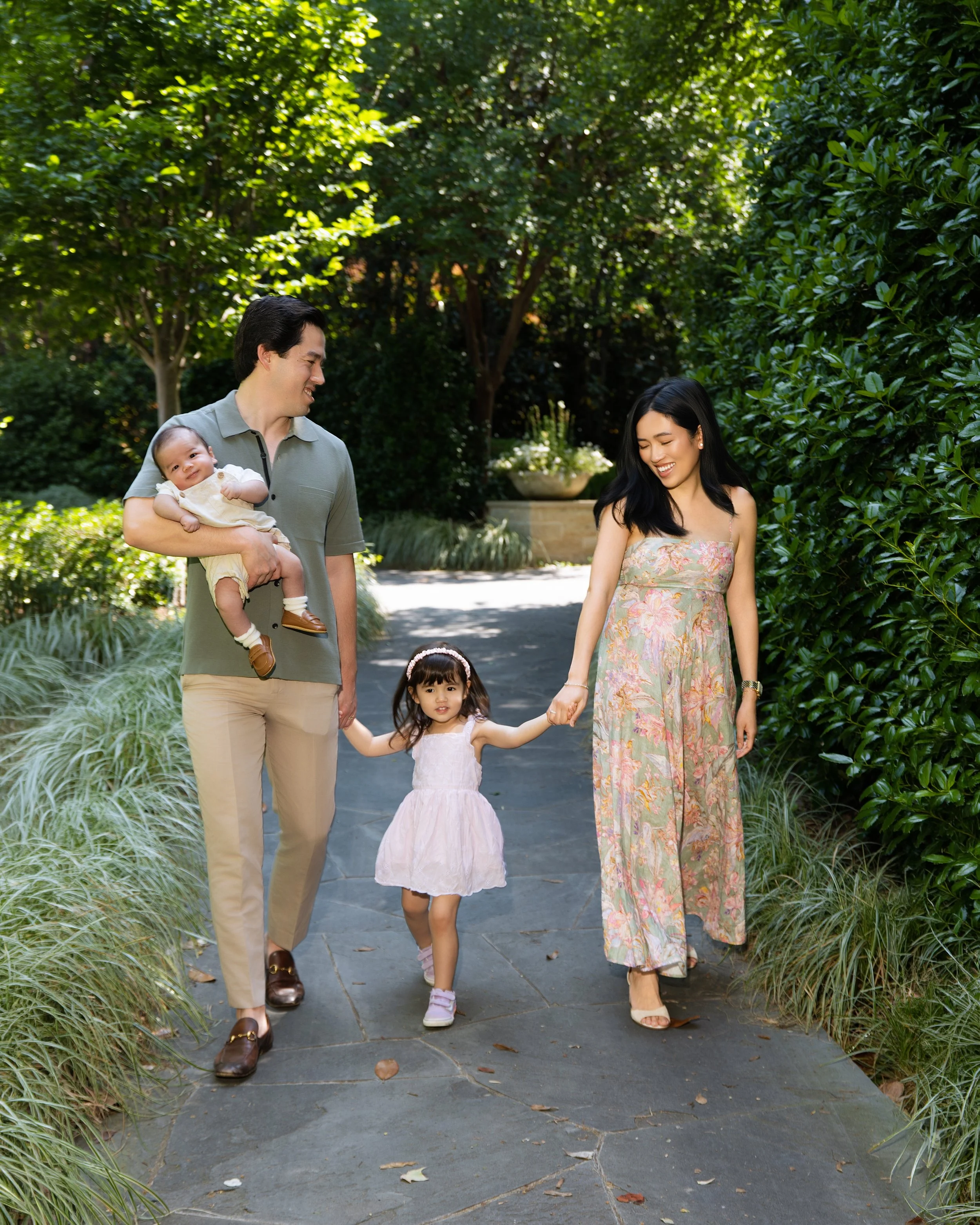 A family of four walking outdoors in a garden. The father, holding a baby, smiling at the mother. The mother, wearing a floral dress, holding hands with a young girl in a white dress. Bright, sunny day with lush green trees and plants.