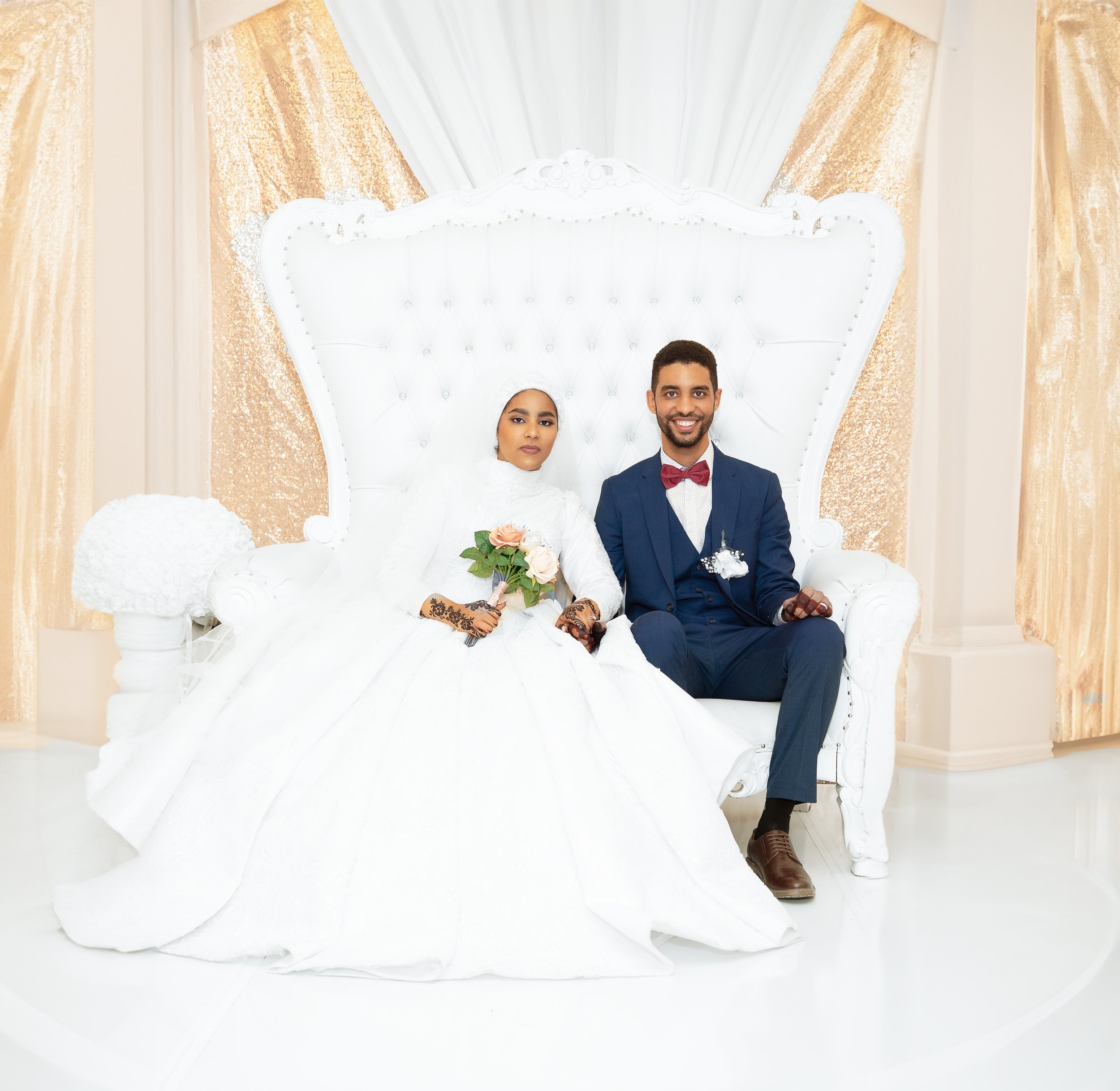 Bride and groom sitting on a white ornate couch; the bride is wearing a white gown and holding a bouquet, the groom in a blue suit with a bow tie.