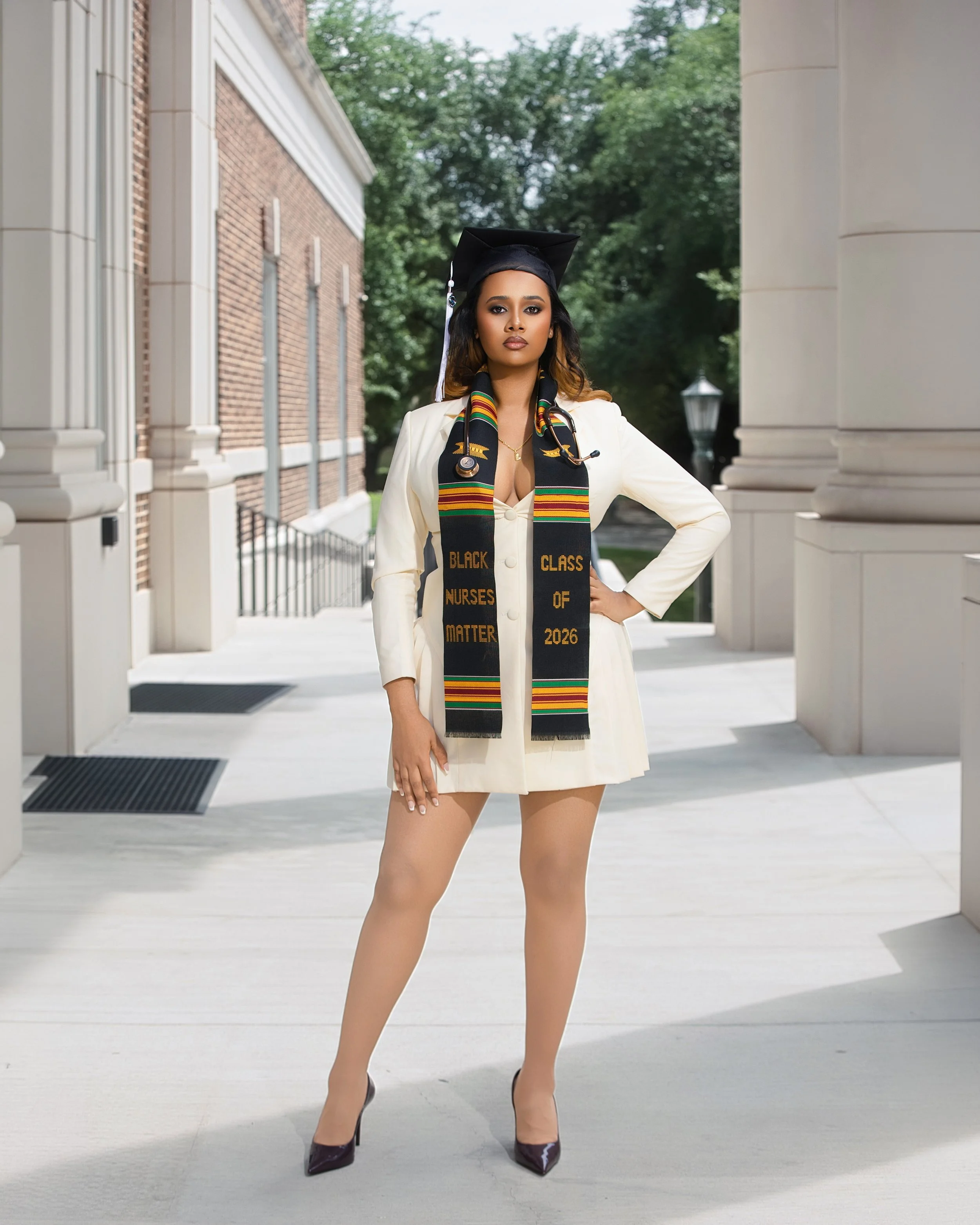 A woman wearing a graduation cap and a cream-colored blazer, with a black stole that says 'Black Nurses Matter' and 'Class of 2026', standing outdoors between two columns with green trees in the background, black high heels, and her hand on her hip.