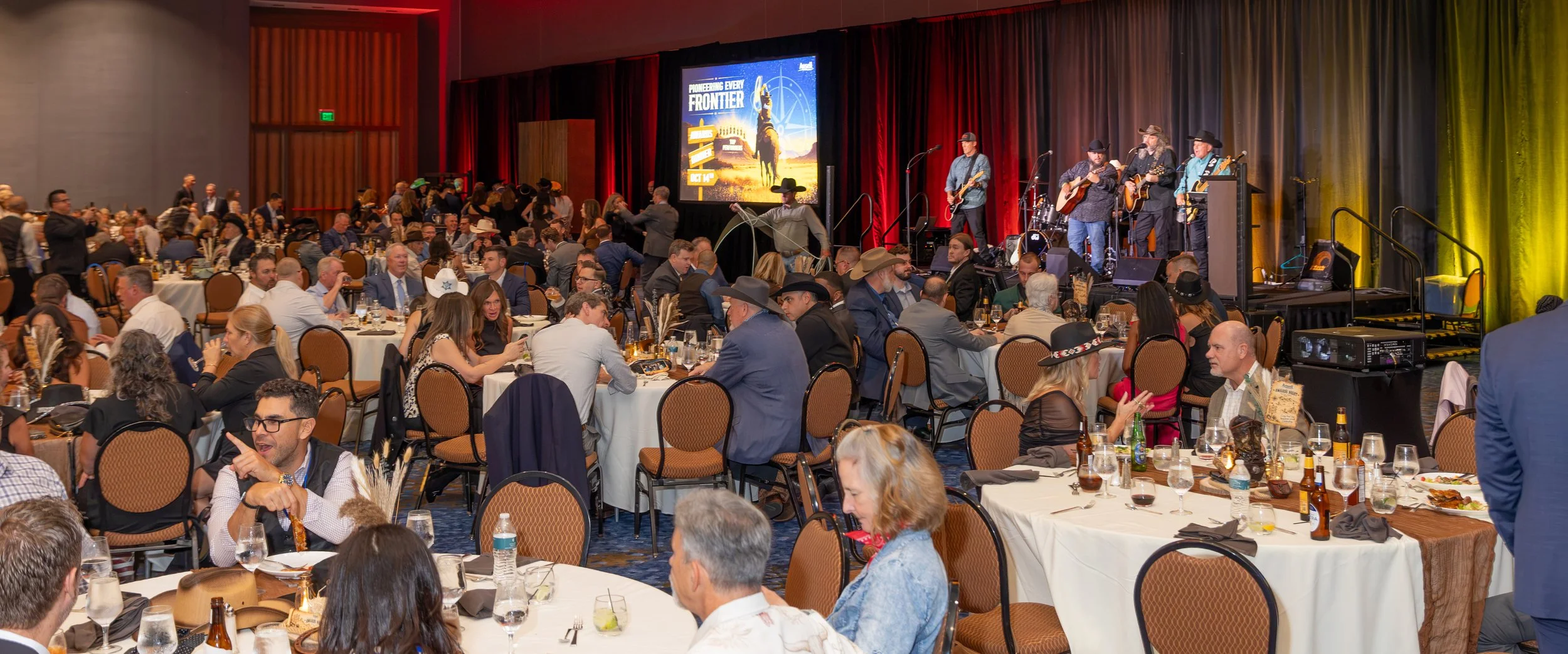 Crowded banquet hall with guests dining while a country band performs onstage beneath red and gold curtains.