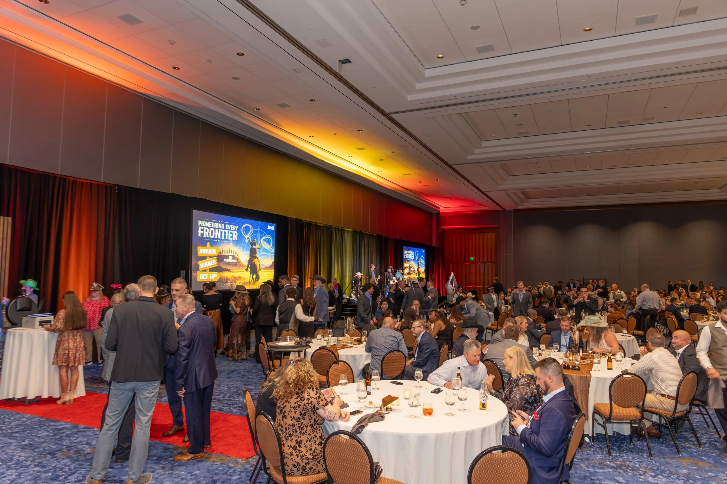 Crowded awards dinner with round tables, a live band, and a large “Pioneering Every Frontier” presentation screen in a brightly lit ballroom