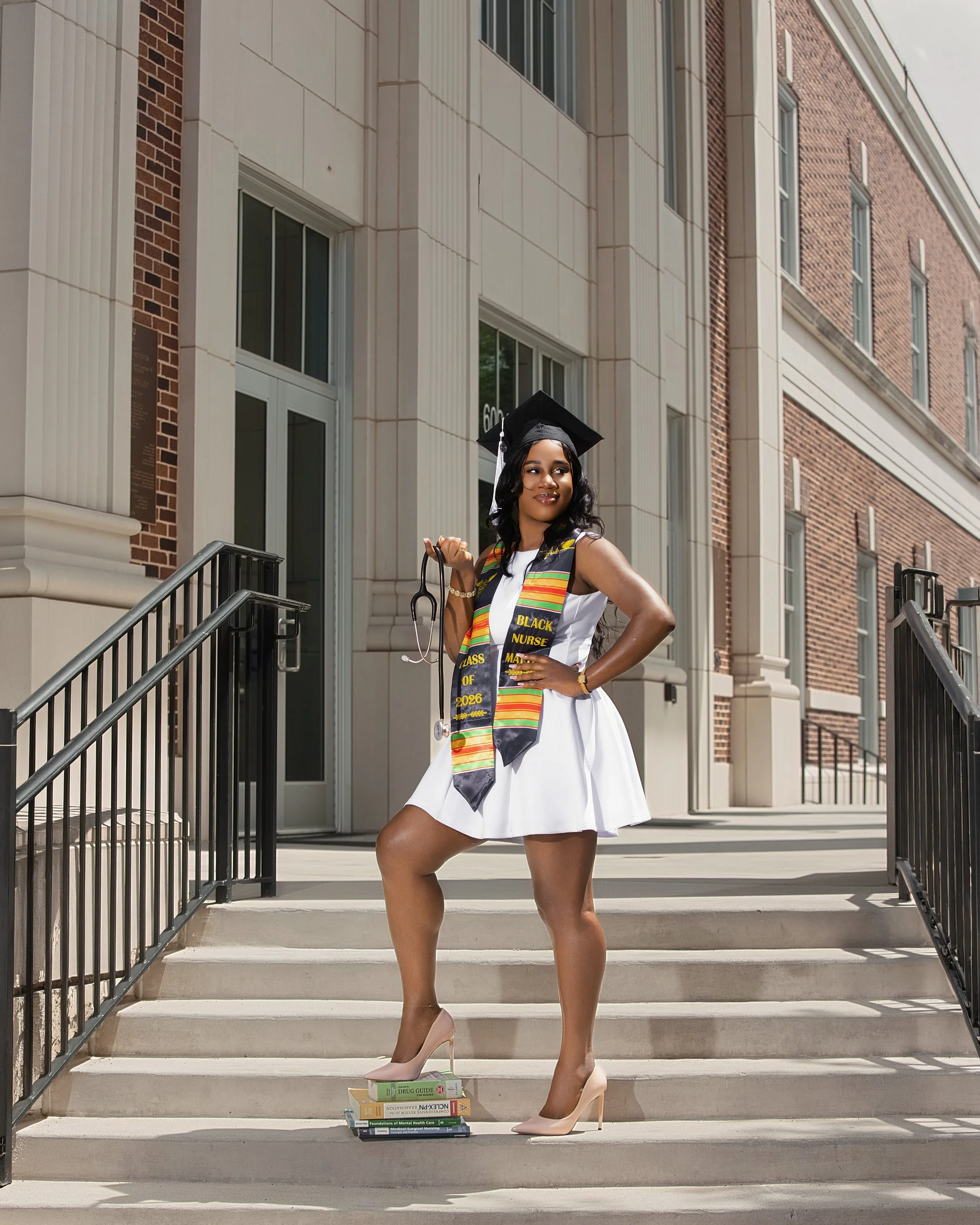 A woman in a graduation cap and gown smiling on the steps outside a building, holding a diploma and stethoscope, with books stacked at her feet. She is wearing high heels and a colorful stole, celebrating her graduation.