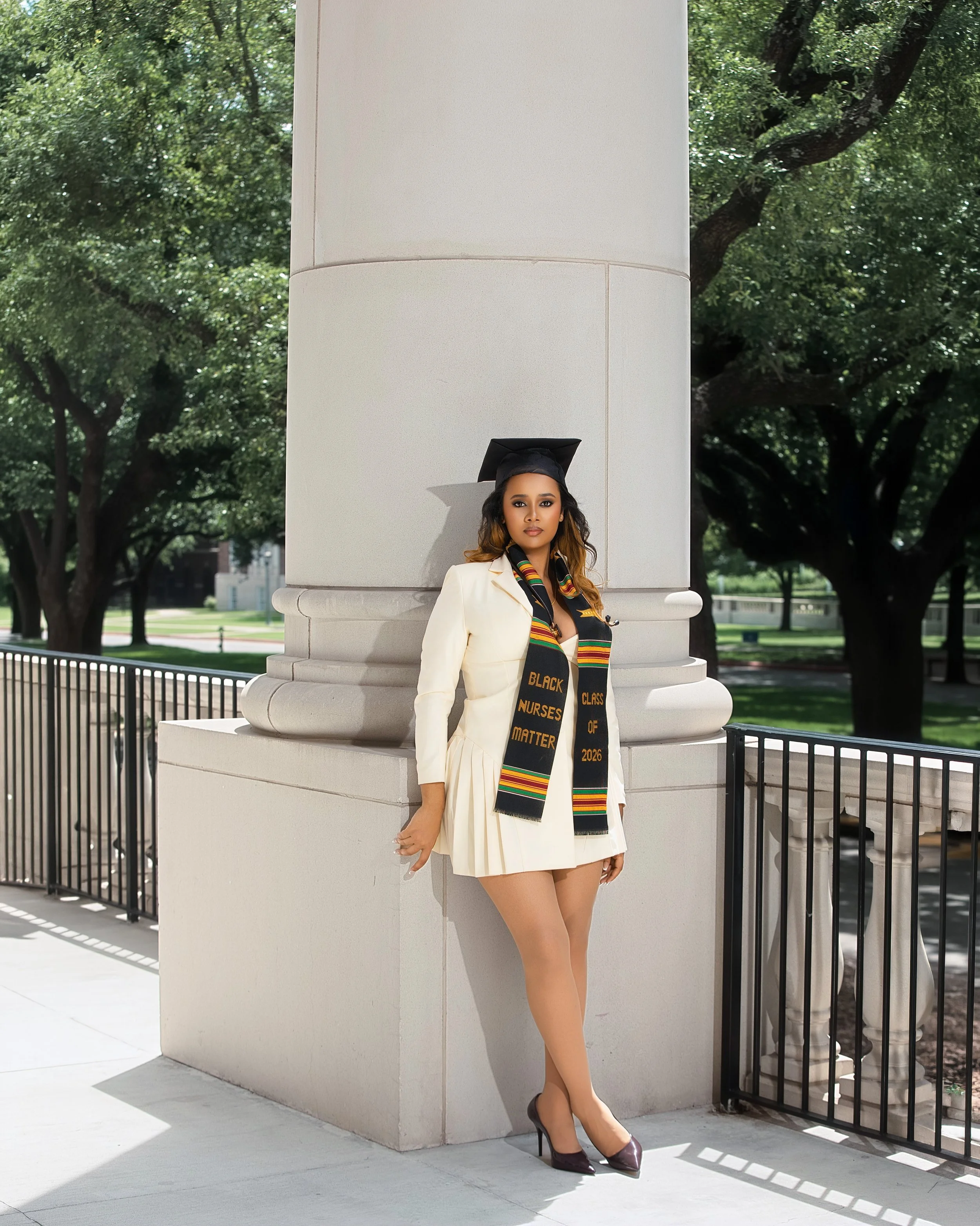 A young woman in a cream-colored blazer and a sorority stole with colorful stripes and the words "Black Nurses Matter" and "Class of 2026" written on it, standing outdoors near a large stone column, wearing a graduation cap and high heels, with trees