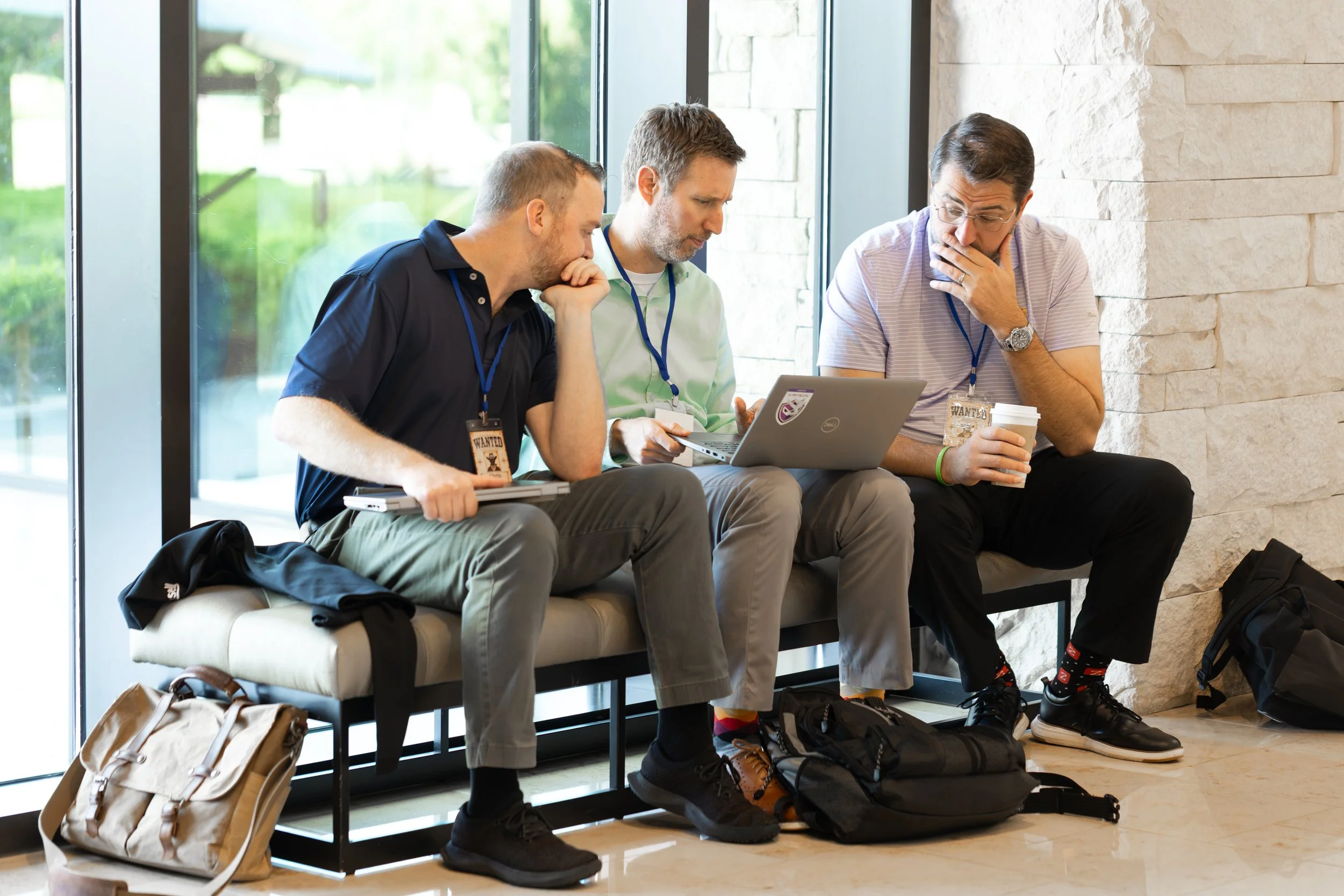 Three men wearing conference badges sit together indoors with laptops and coffee, discussing work near a window.
