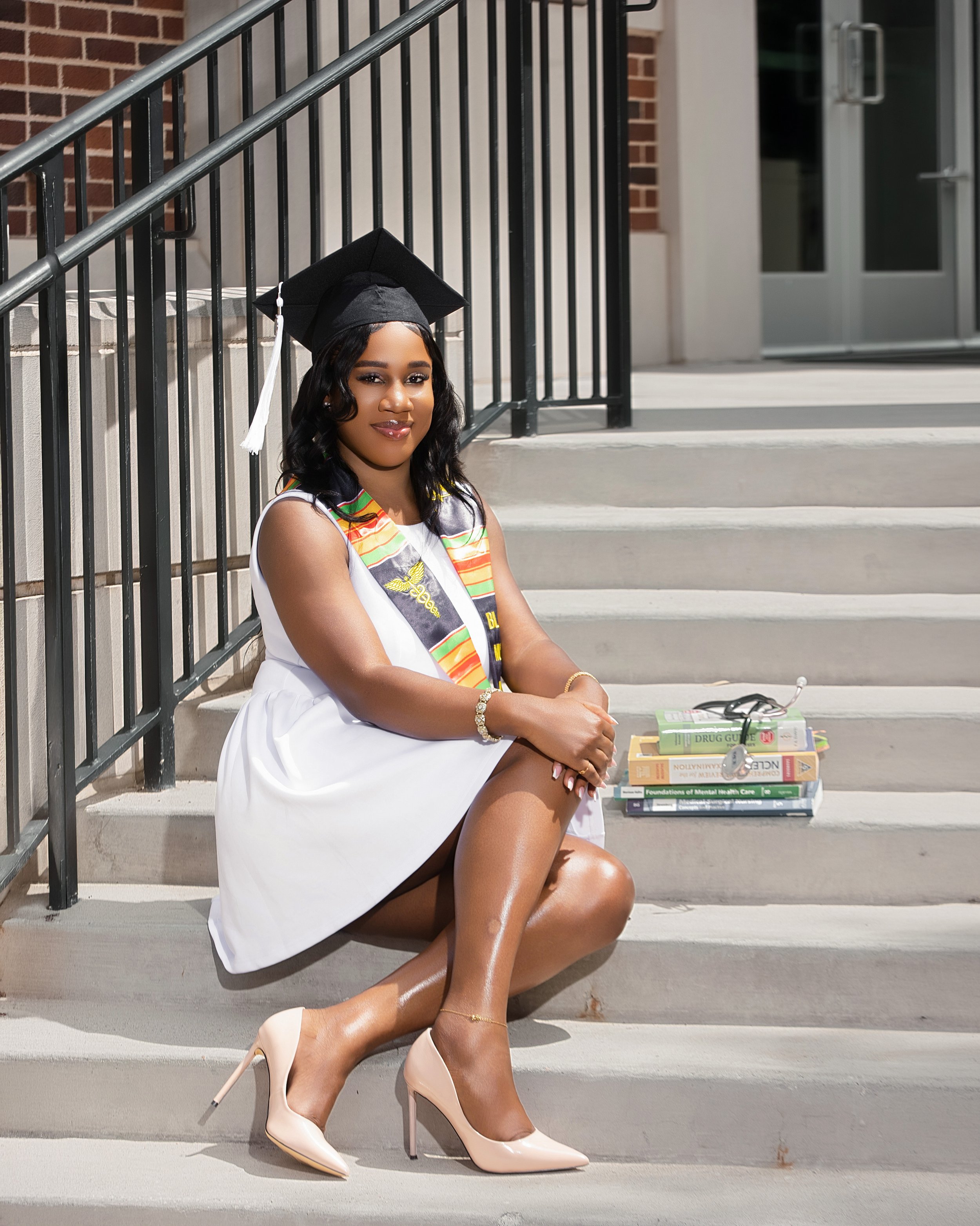 A woman in a white dress and beige high heels is sitting on steps outside a building, wearing a graduation cap and stole. She is smiling and looking at the camera, with books on mental health and drug guides near her on the steps.