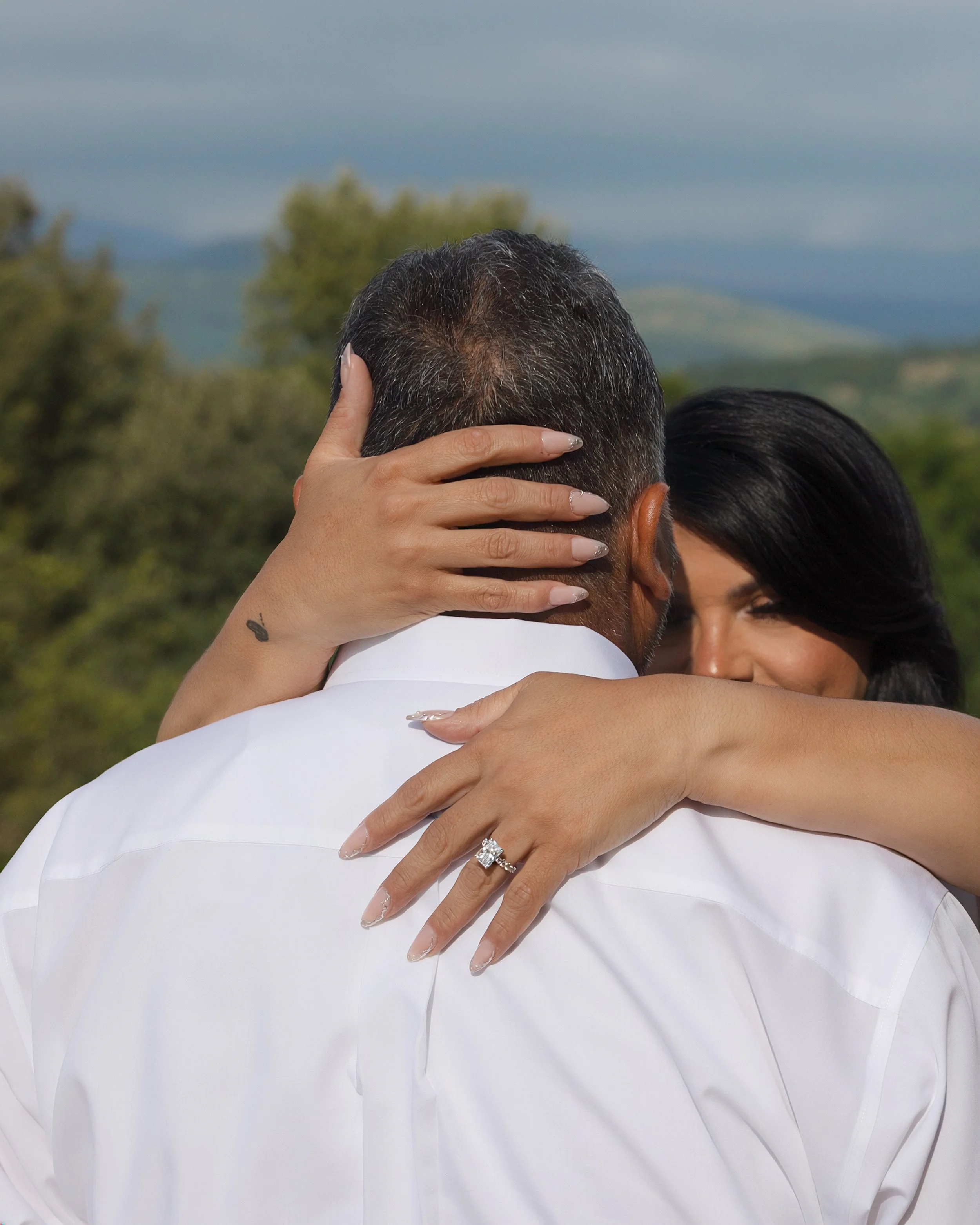 Close-up of a couple embracing outdoors during daytime, with focus on their heads and arms, and a scenic background of trees and hills.