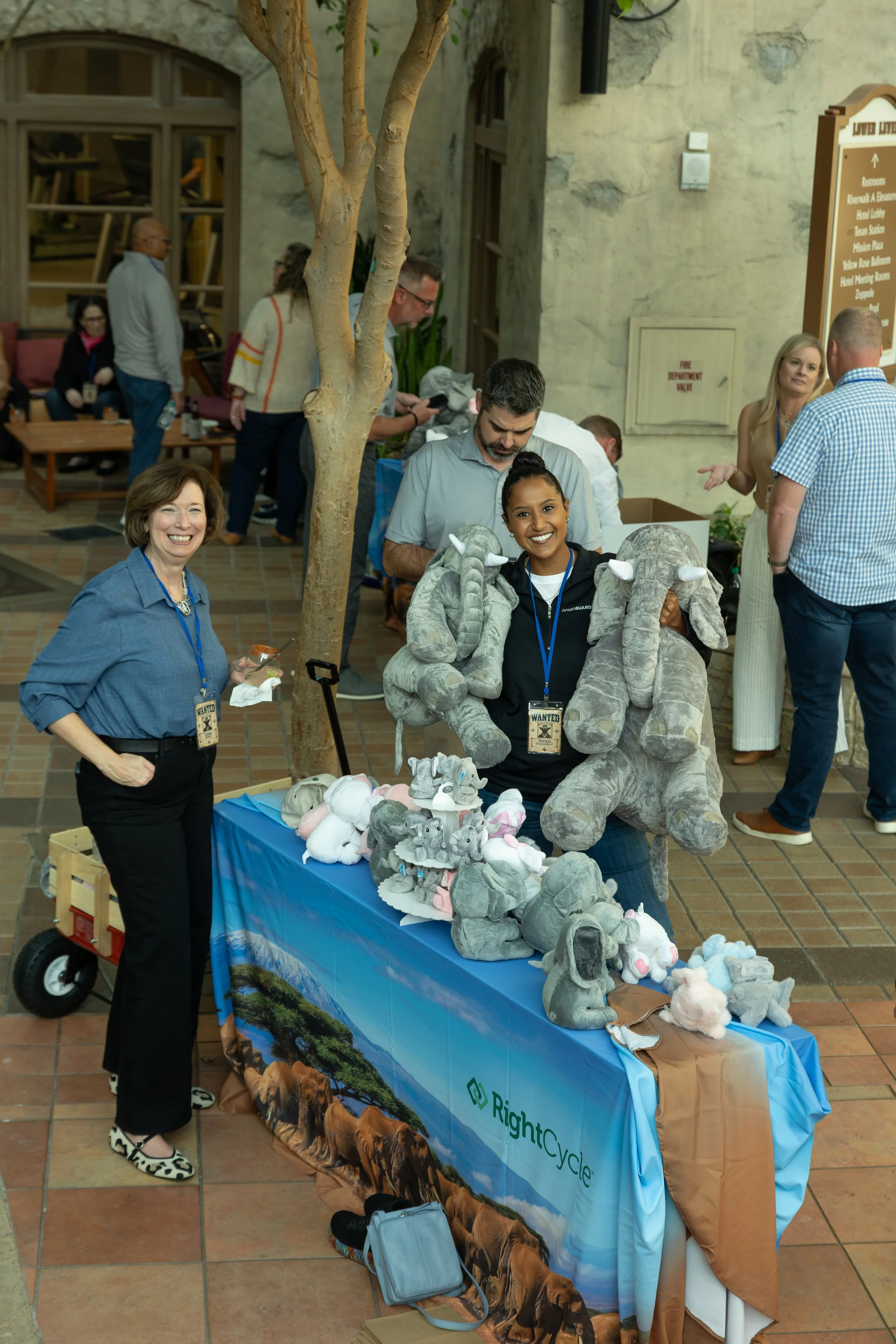 People gather at a RightCycle recycling event table displaying plush elephant toys inside a building.