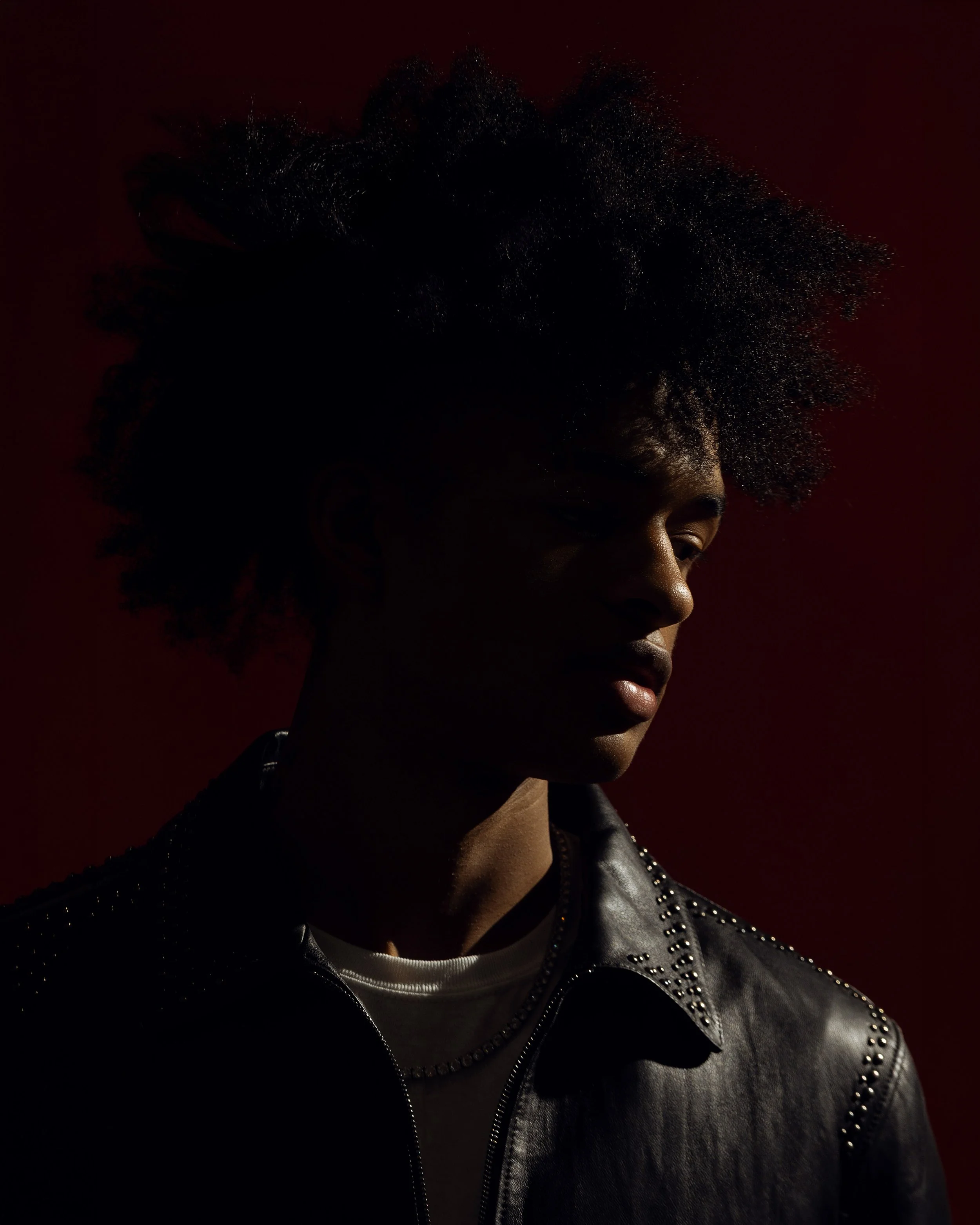 A person with curly hair, wearing a dark leather jacket with silver studs, in profile against a dark red background.