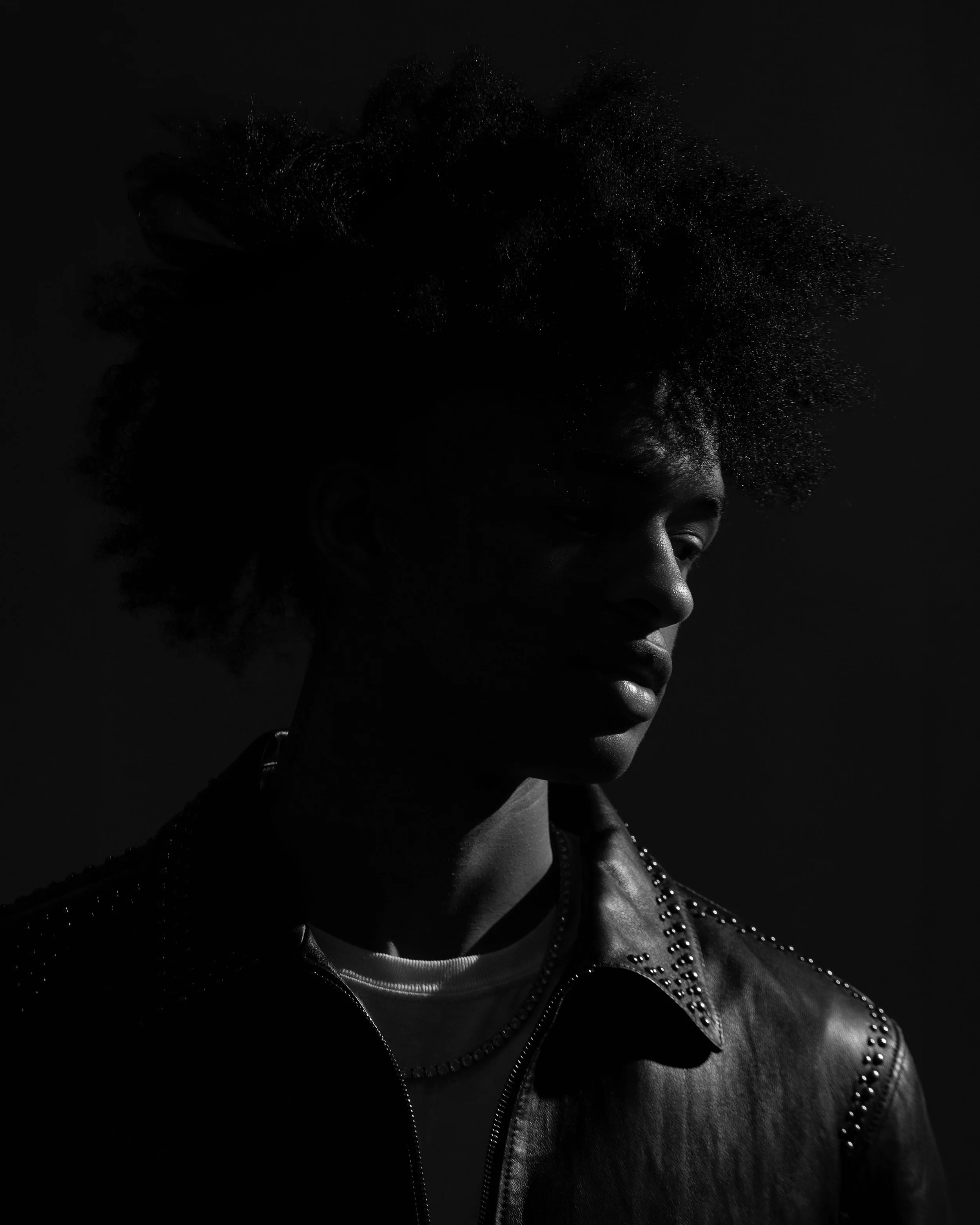 A black and white portrait of a person with textured, curly hair, wearing a studded leather jacket, looking down with a serious expression.