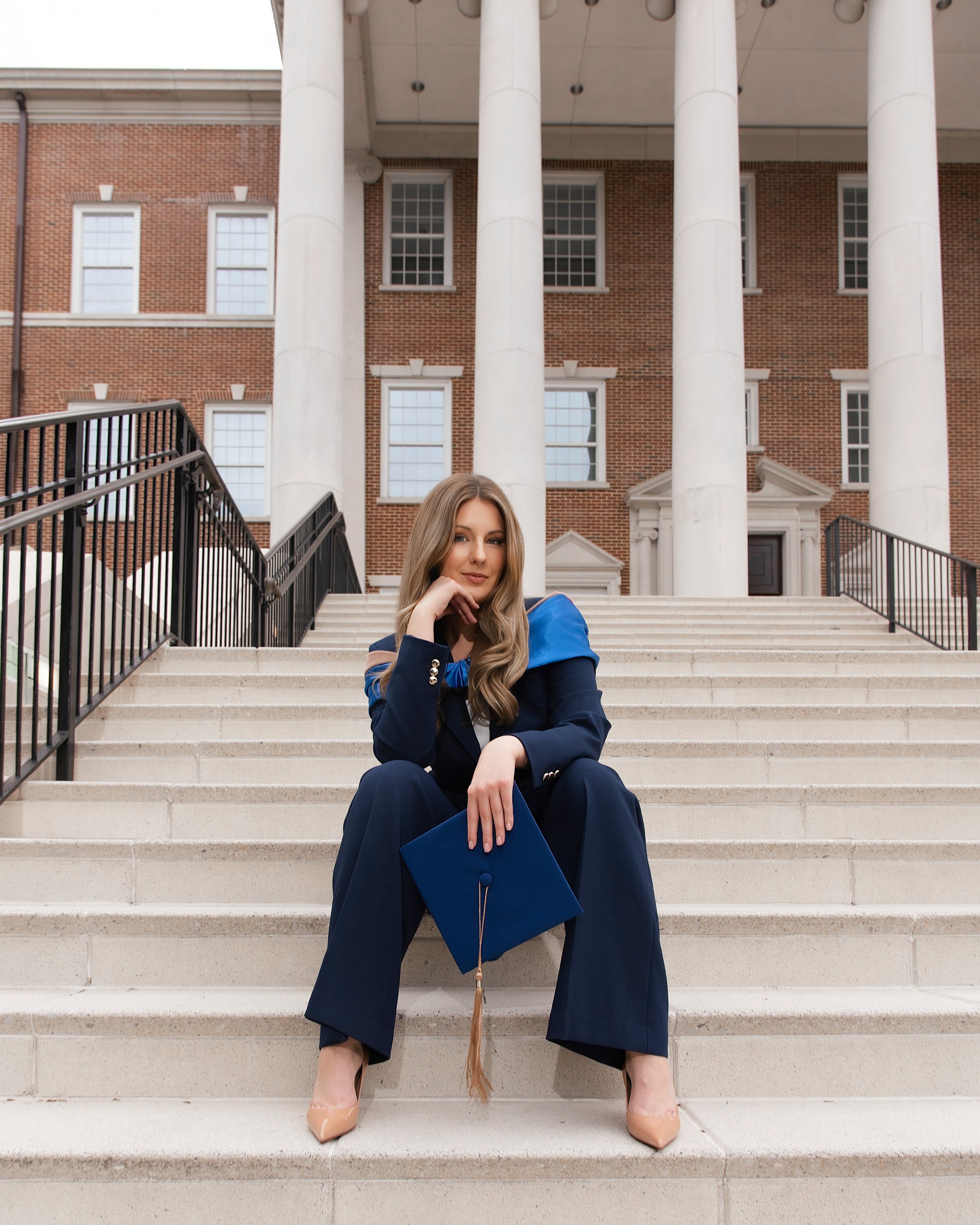 Young woman in graduation cap and gown sitting on stone steps outside a brick building with tall white columns.