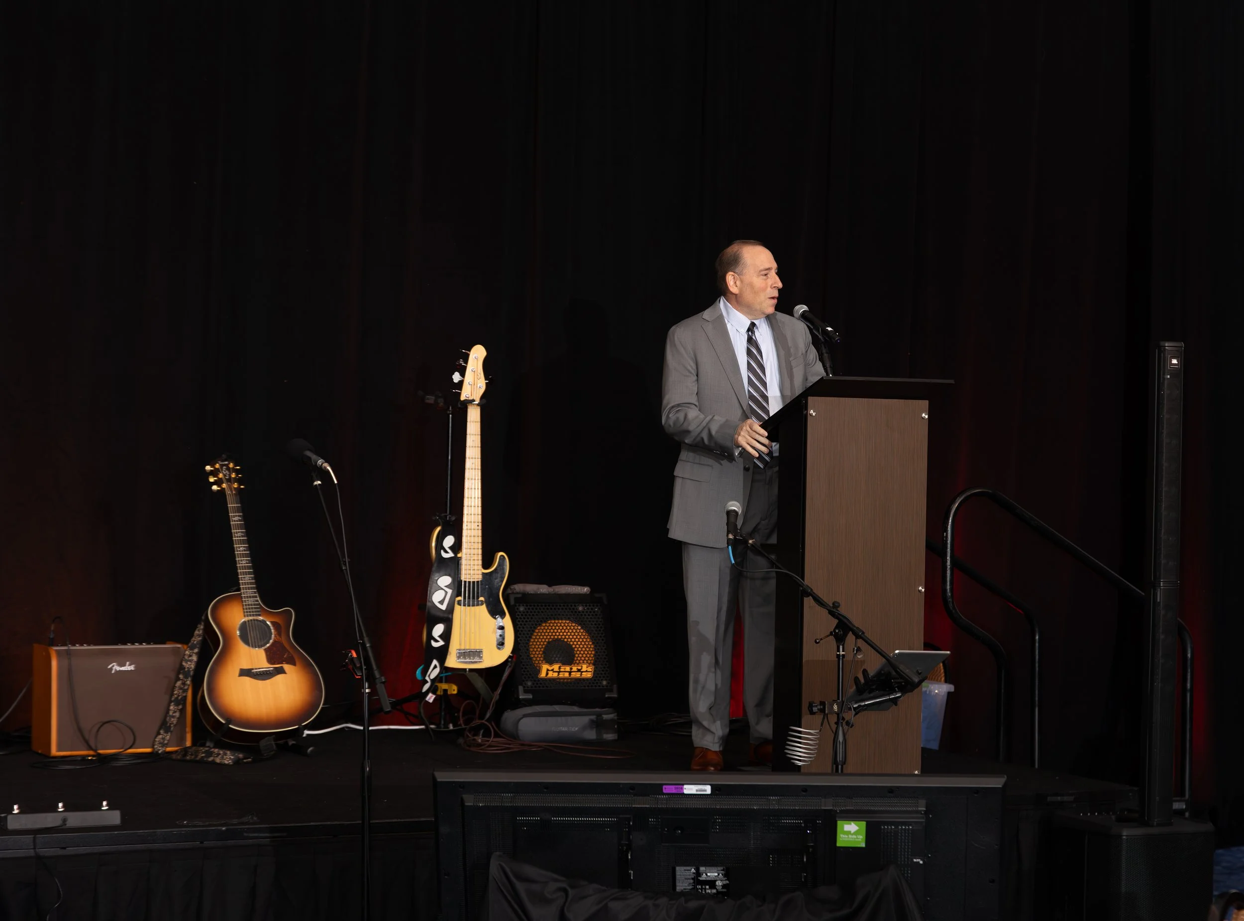 Man in a gray suit speaking at a podium on a stage with guitars, microphones, and audio equipment.