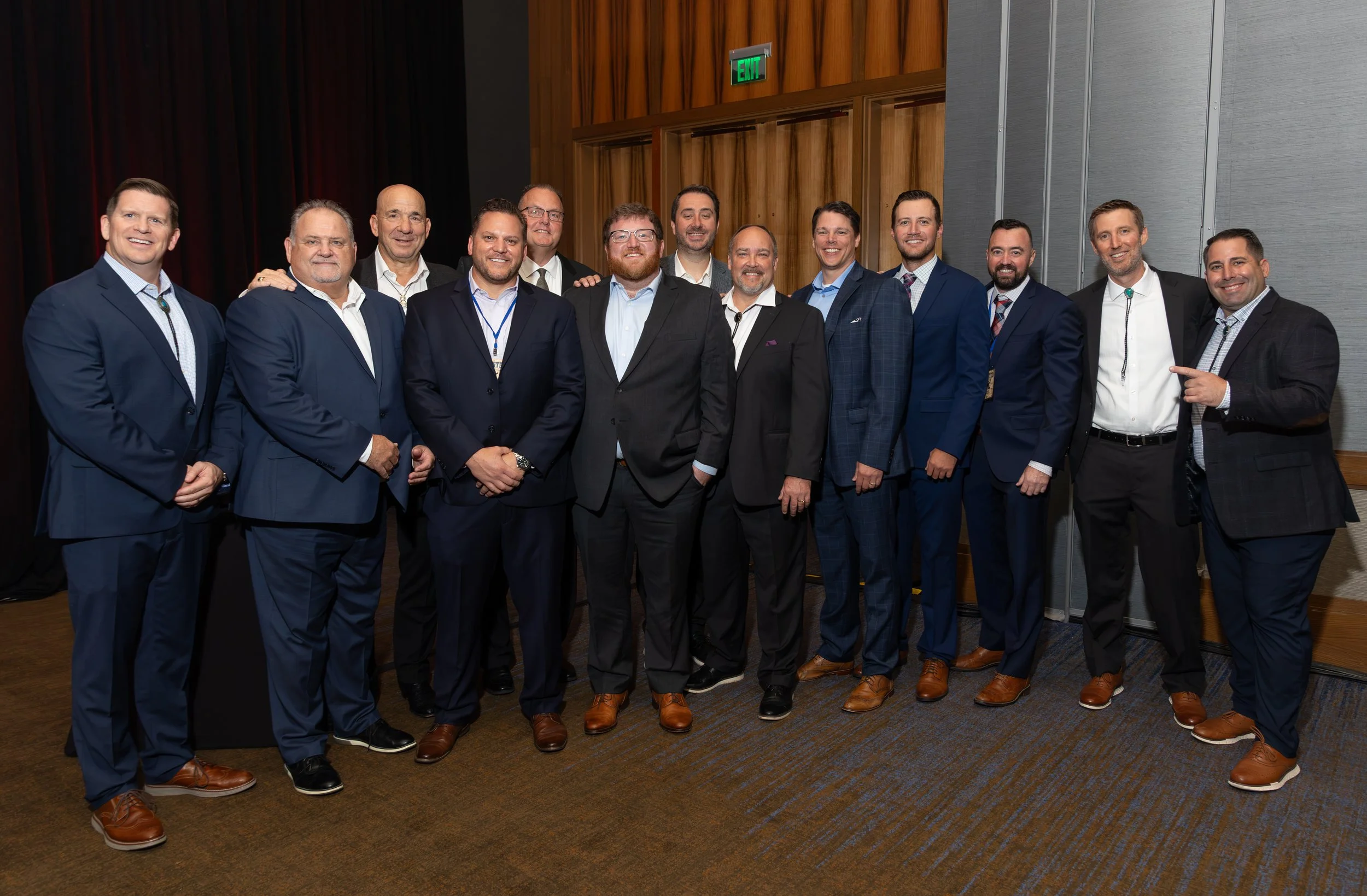 Group of men in suits posing for a photo in a wood-paneled event hall