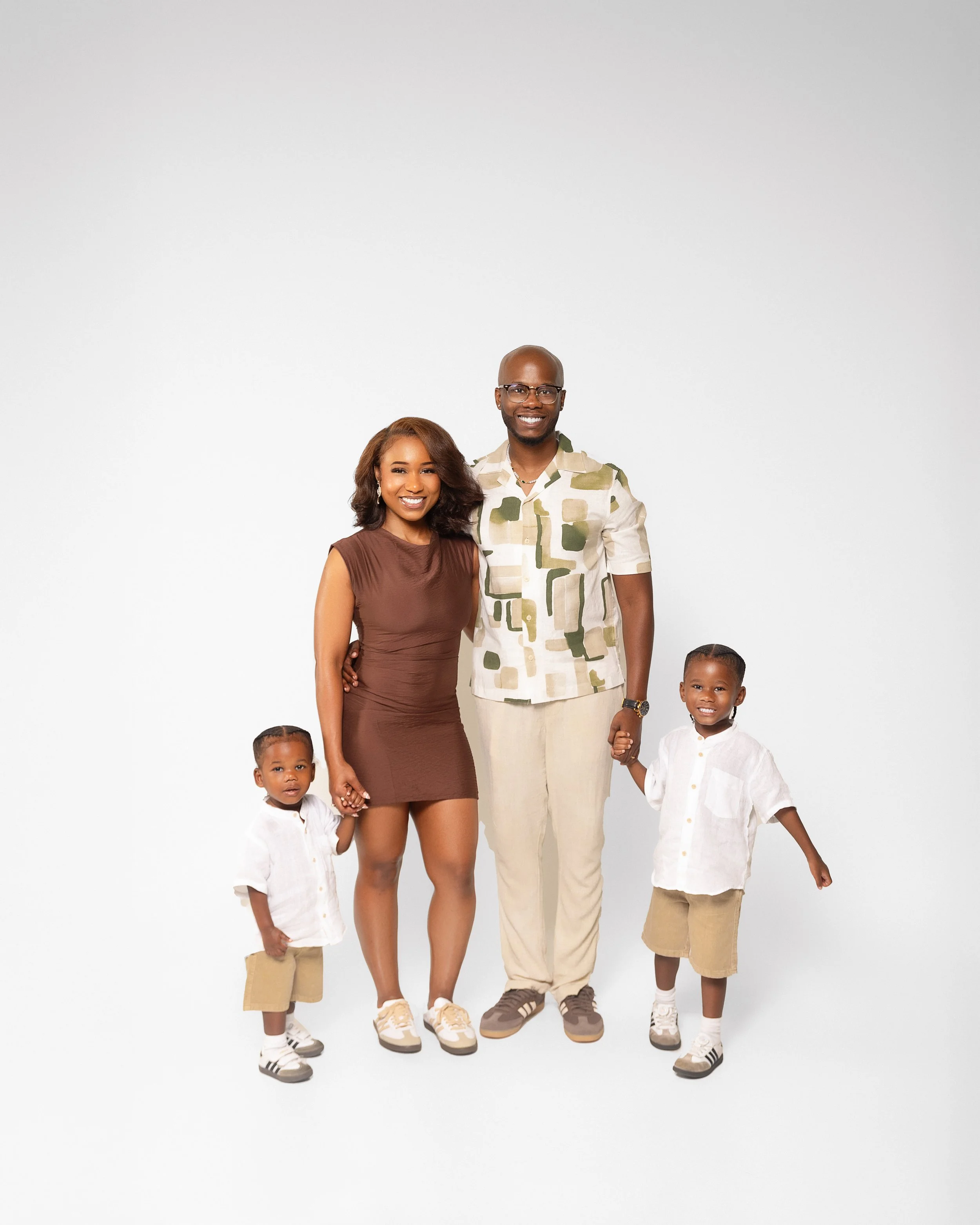 A happy African American family of four standing together on a white background. The mother has shoulder-length brown hair and is wearing a sleeveless brown dress. The father, bald and wearing glasses, is dressed in a patterned short-sleeve shirt and