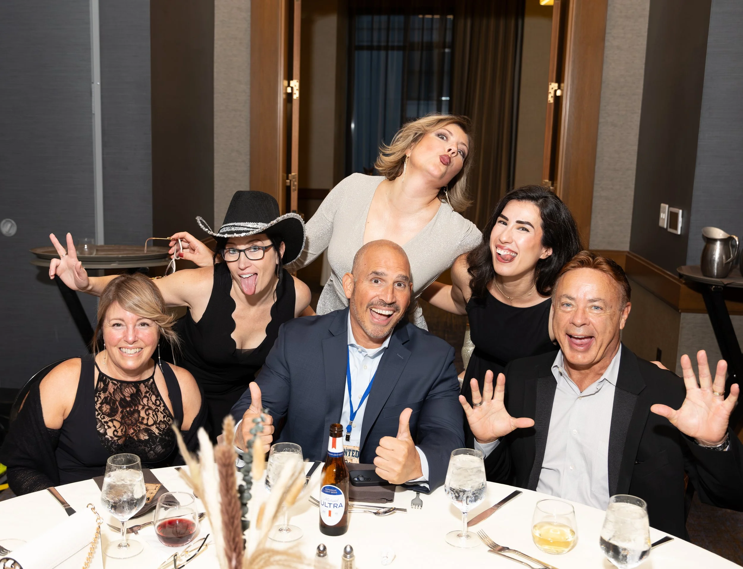 Group of six friends posing playfully at a restaurant dinner table, with drinks and a beer bottle