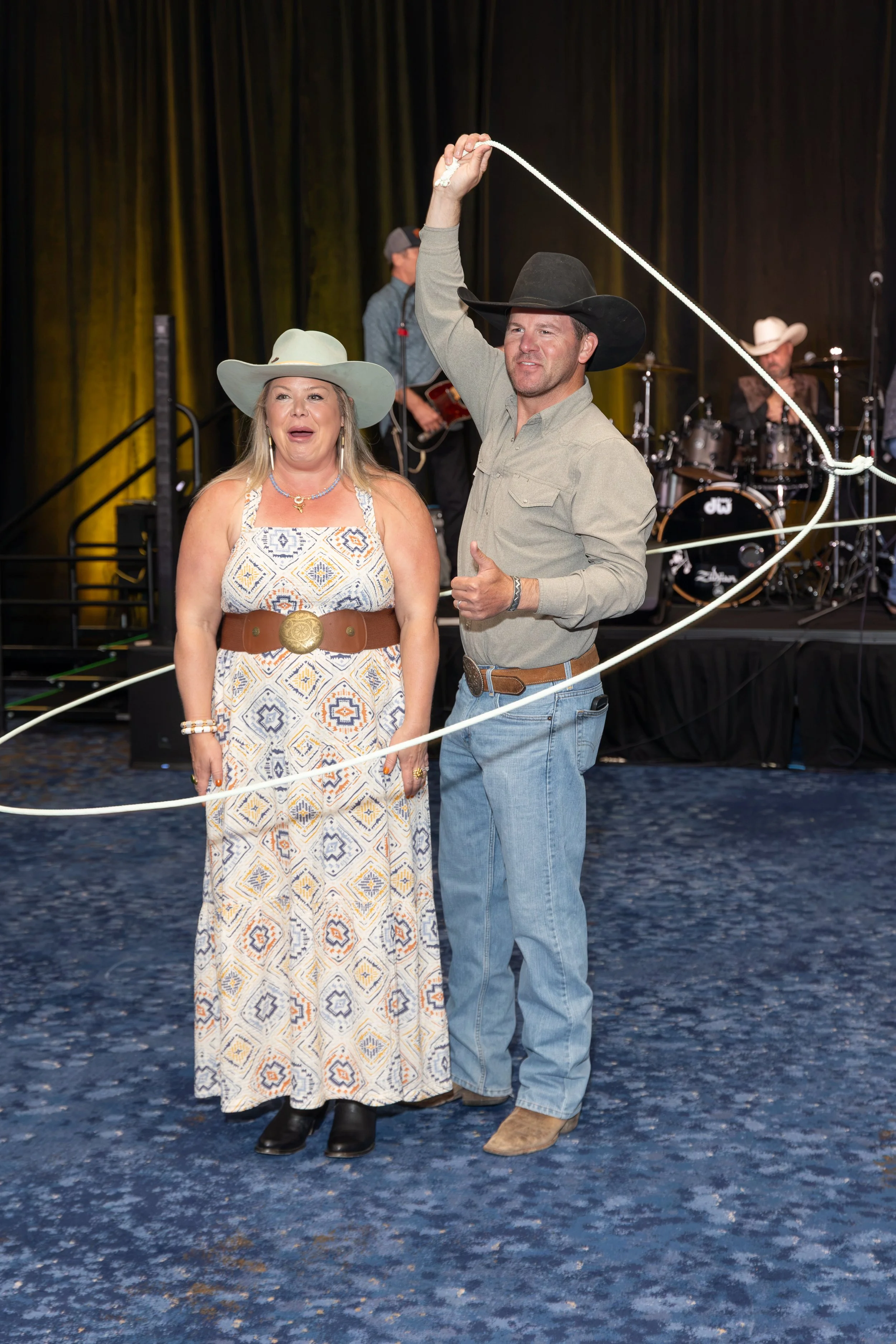 Man in a black cowboy hat holding a white lasso beside a woman in a cowboy hat on a country music stage with a band.