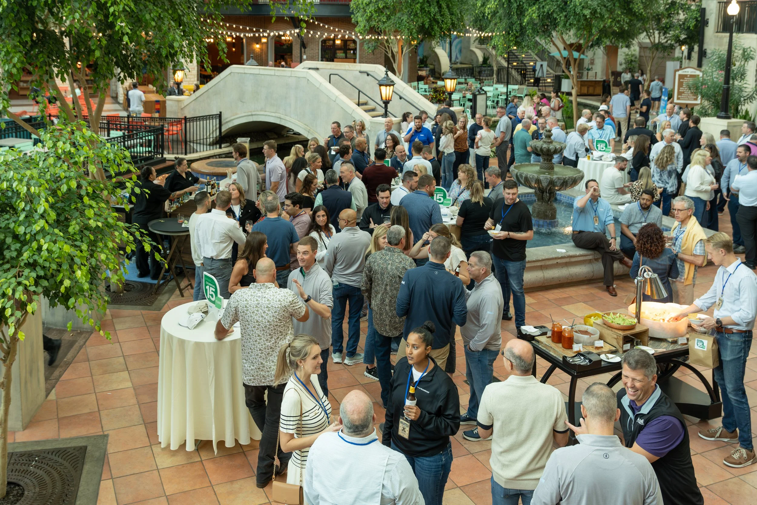 Crowded indoor courtyard reception with guests, buffet tables, string lights, greenery, a fountain, and arched footbridge
