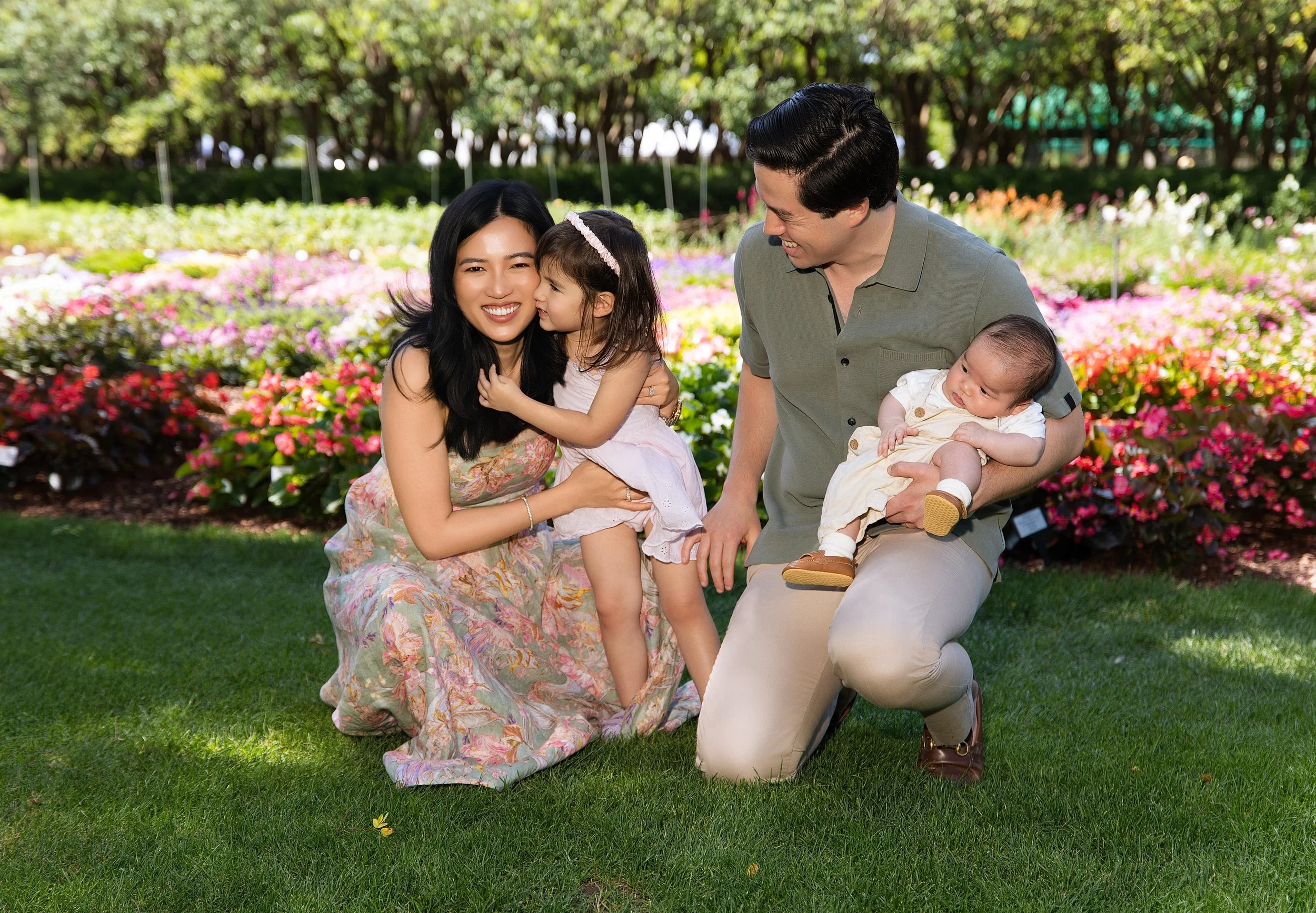 A family of four, including a woman, a man, a young girl, and a baby, enjoying a moment together in a garden with colorful flowers and green trees in the background.