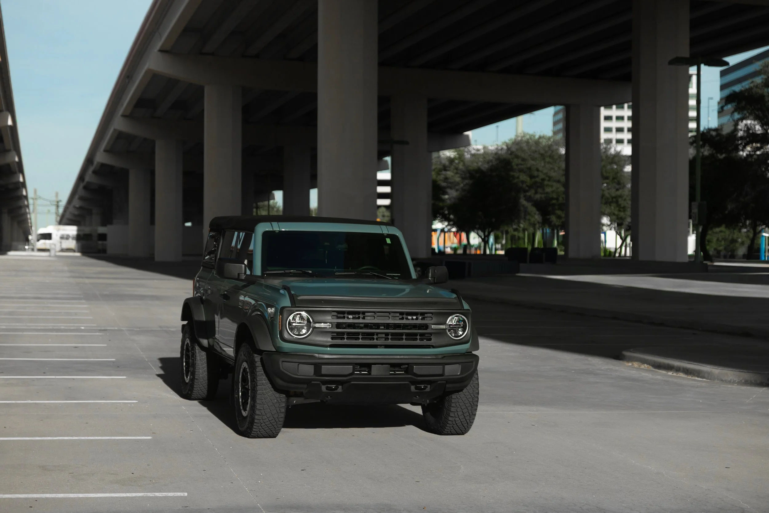 A small dark green SUV parked alone in an urban parking lot under a bridge with tall buildings and trees in the background.