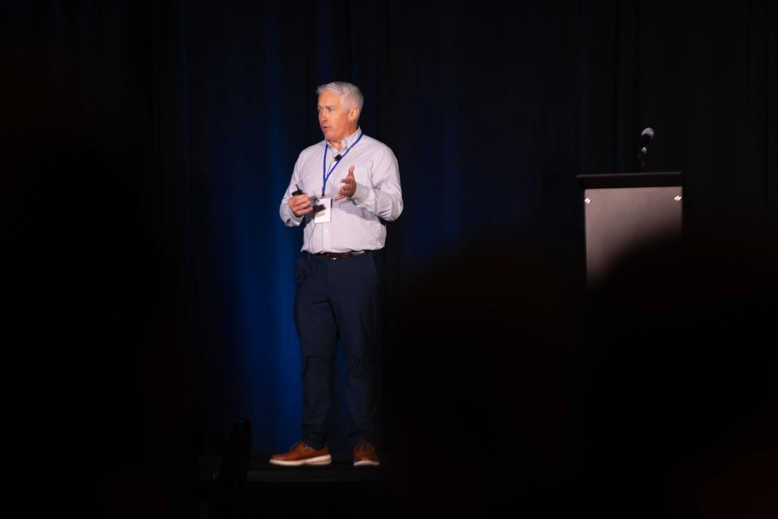 Gray-haired man giving a presentation on a dark stage beside a podium and microphone