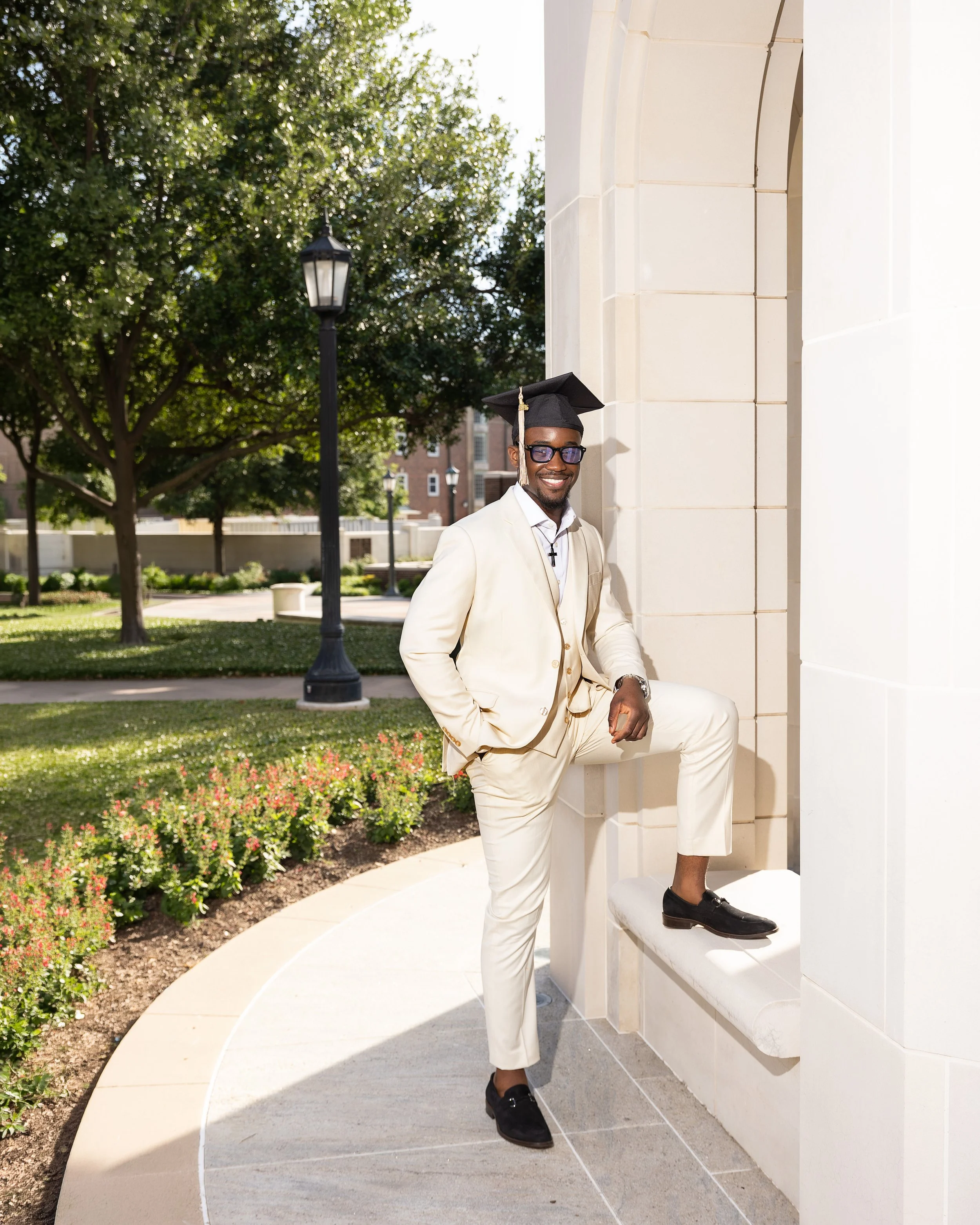 A man dressed in a light cream suit with a white shirt and black shoes, wearing a black graduation cap, glasses, and a cross necklace, posing outdoors next to a classical building with a large archway. He is standing with one foot on a ledge and smil