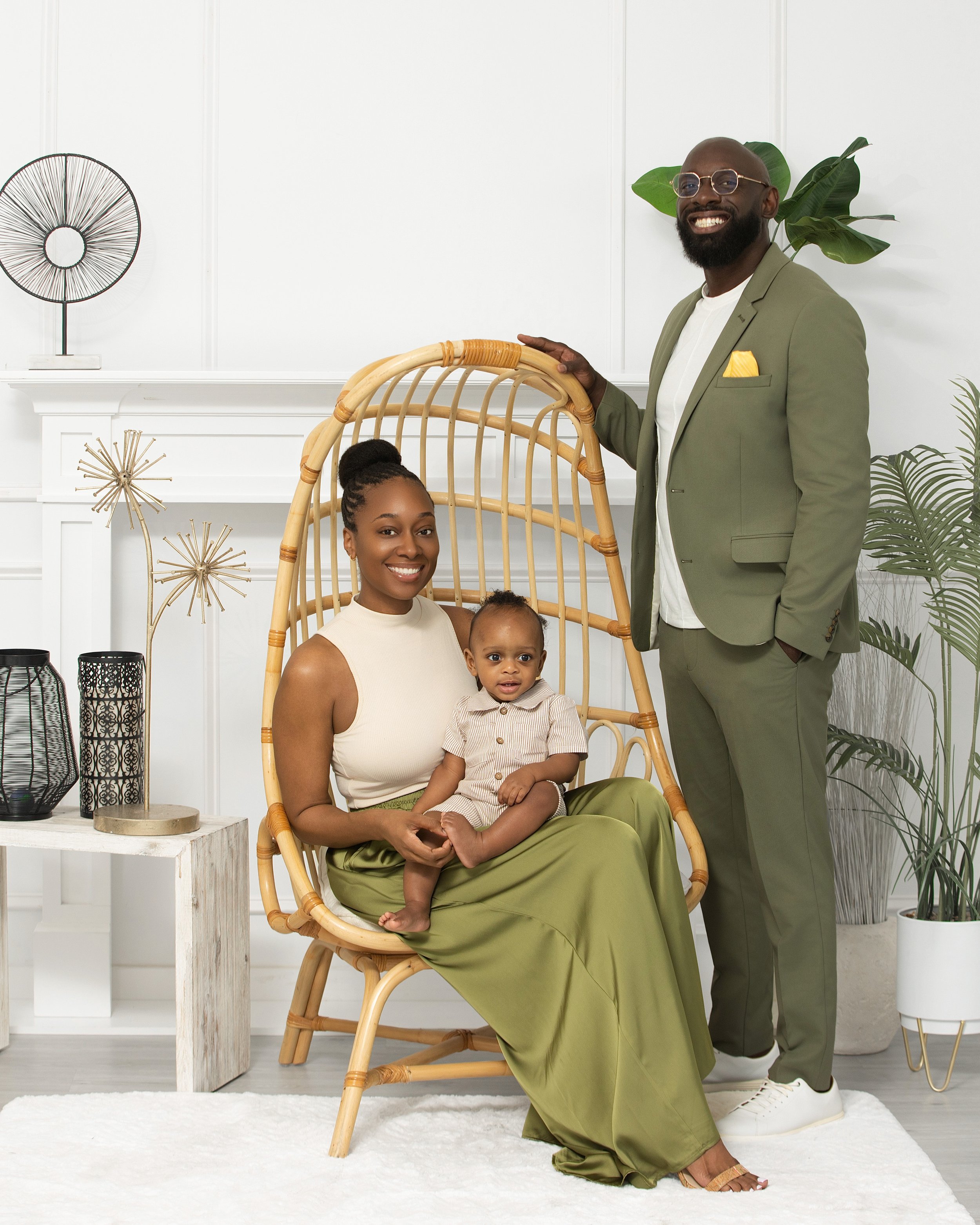 A woman sitting on a chair with a young child on her lap, and a man standing behind them, all smiling in a modern, bright room with plants and decorative items.