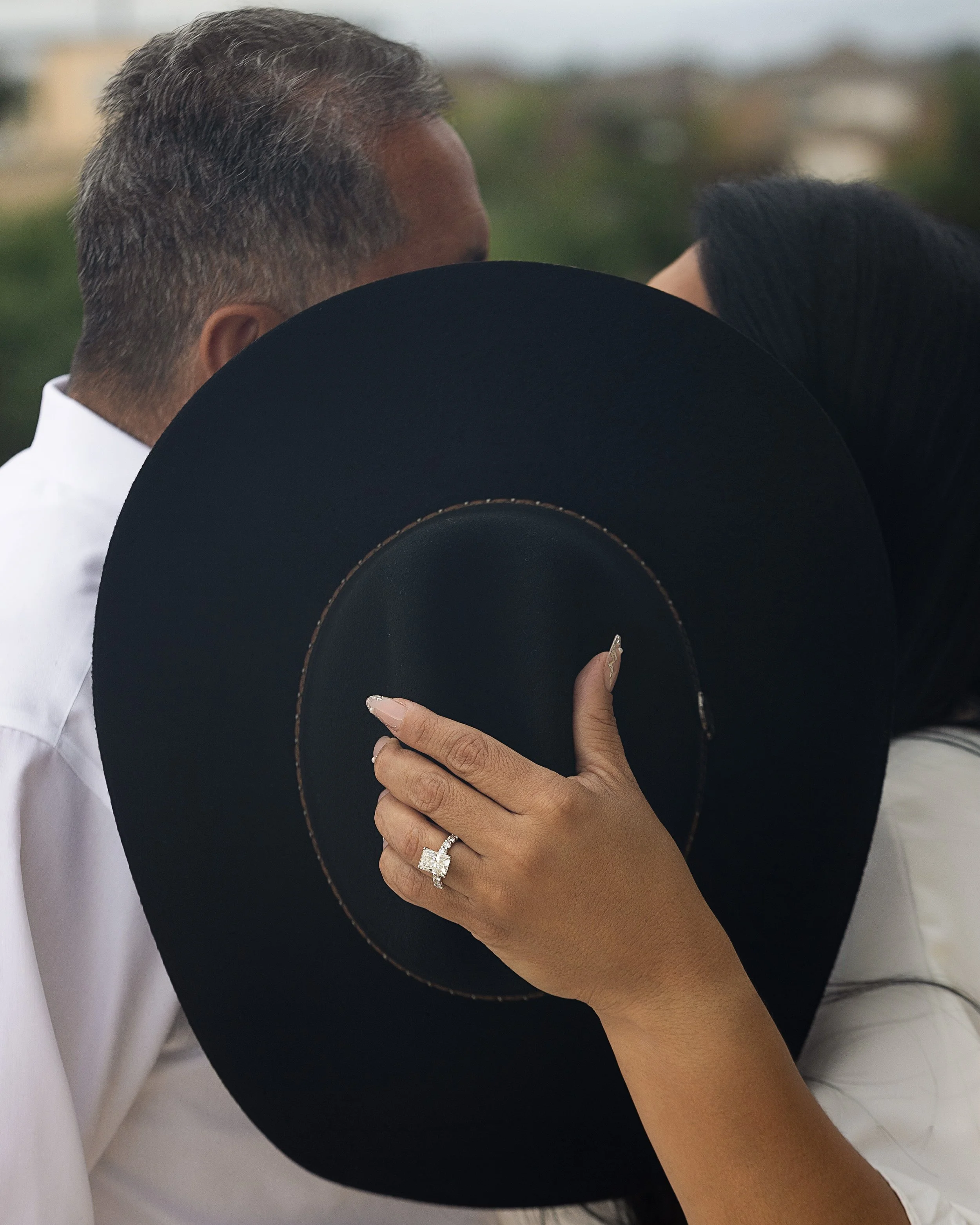 A woman is holding a black hat with a gold band. She is wearing a large square-cut diamond ring on her ring finger. Her arm and hand are in focus, with her nails painted in a light color. The background shows part of a person's head and a white shirt