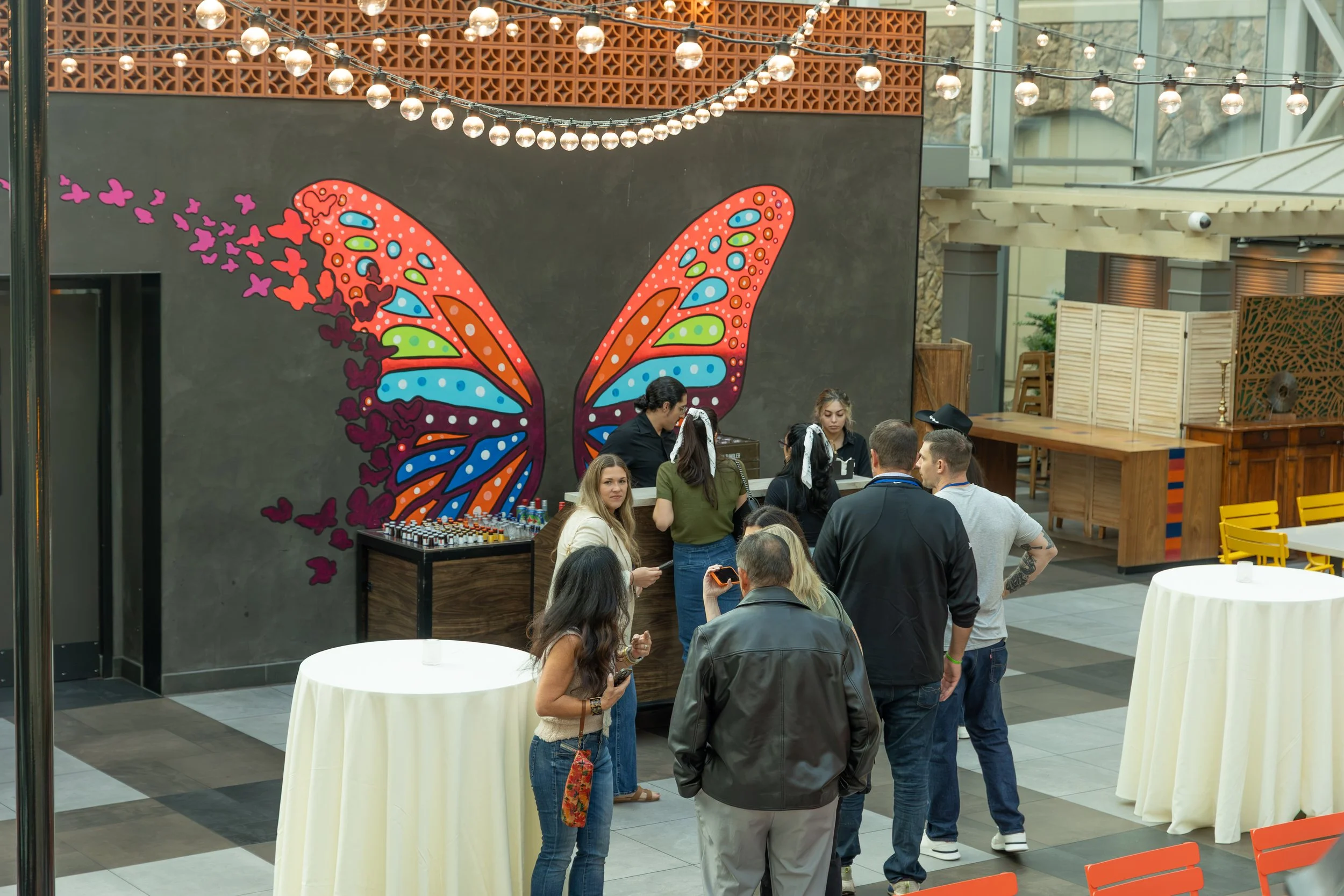 Visitors gather at an event beneath string lights in front of a colorful butterfly mural and refreshment table.