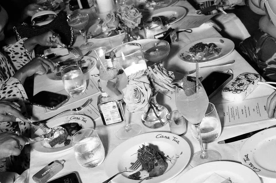 A table set for dinner at a restaurant with plates of food, glasses of water and cocktails, and various personal items, including a phone and a wallet.