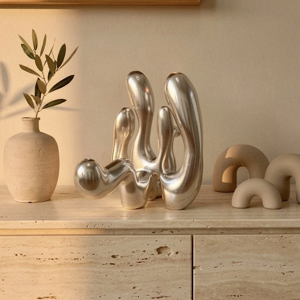 Decorative arrangement on a light-colored wooden surface with a beige vase holding green leafy sprigs, a shiny metallic abstract sculpture, and three small beige arch-shaped sculptures.